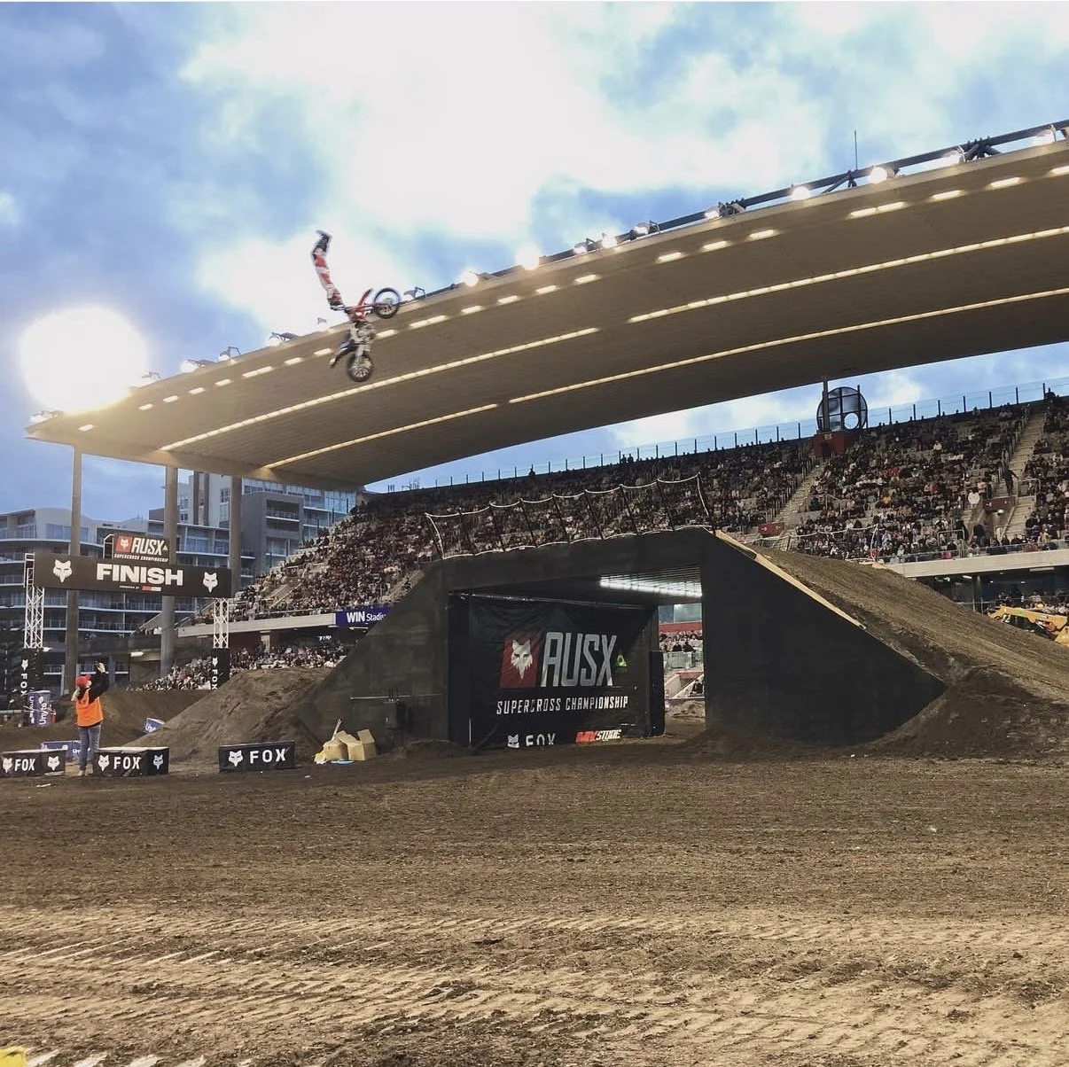 A motocross rider performing a mid-air trick during a race at the AUSX Supercross Championship with spectators in the background.