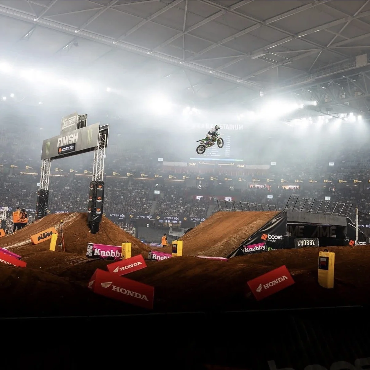 A motocross rider mid-air over a dirt jump at an indoor stadium with a large crowd, finish line arch, and sponsorship banners.