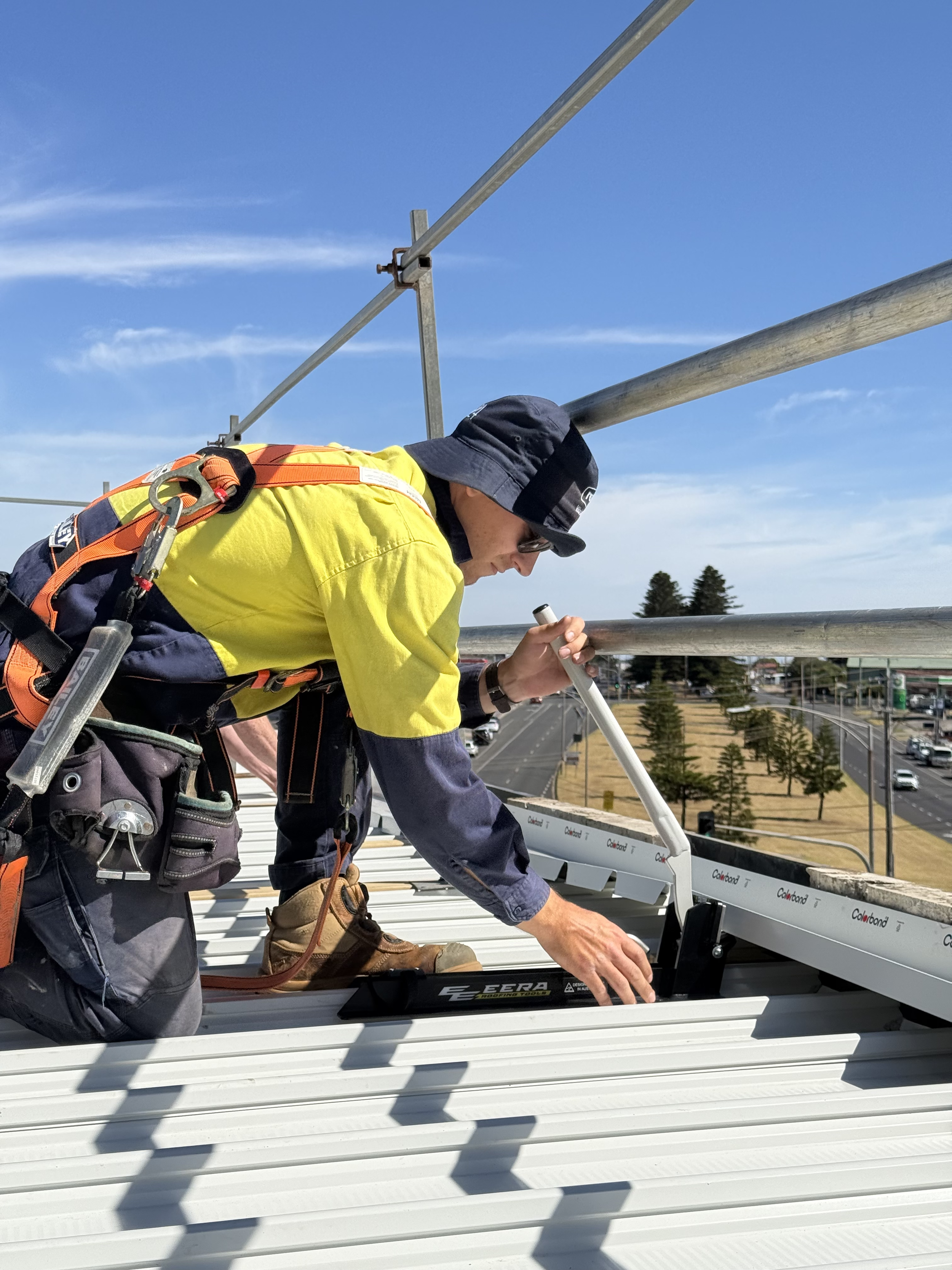 Construction worker wearing safety gear installs or inspects roof panels on a building with a city view in the background.