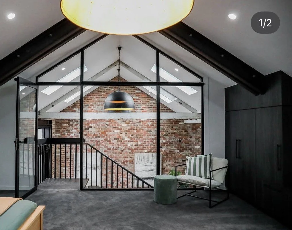 Modern loft interior with a brick wall, skylights, black framing, and a cozy seating area with a striped pillow and green ottoman.