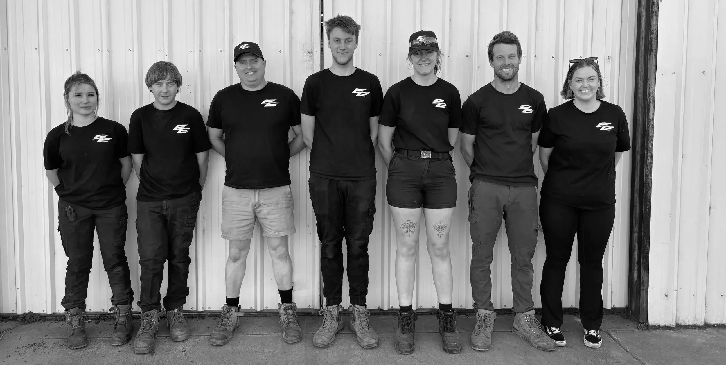Group of seven people standing side by side against a corrugated metal wall, smiling, in casual black t-shirts and pants or shorts, some with tattoos and sunglasses.