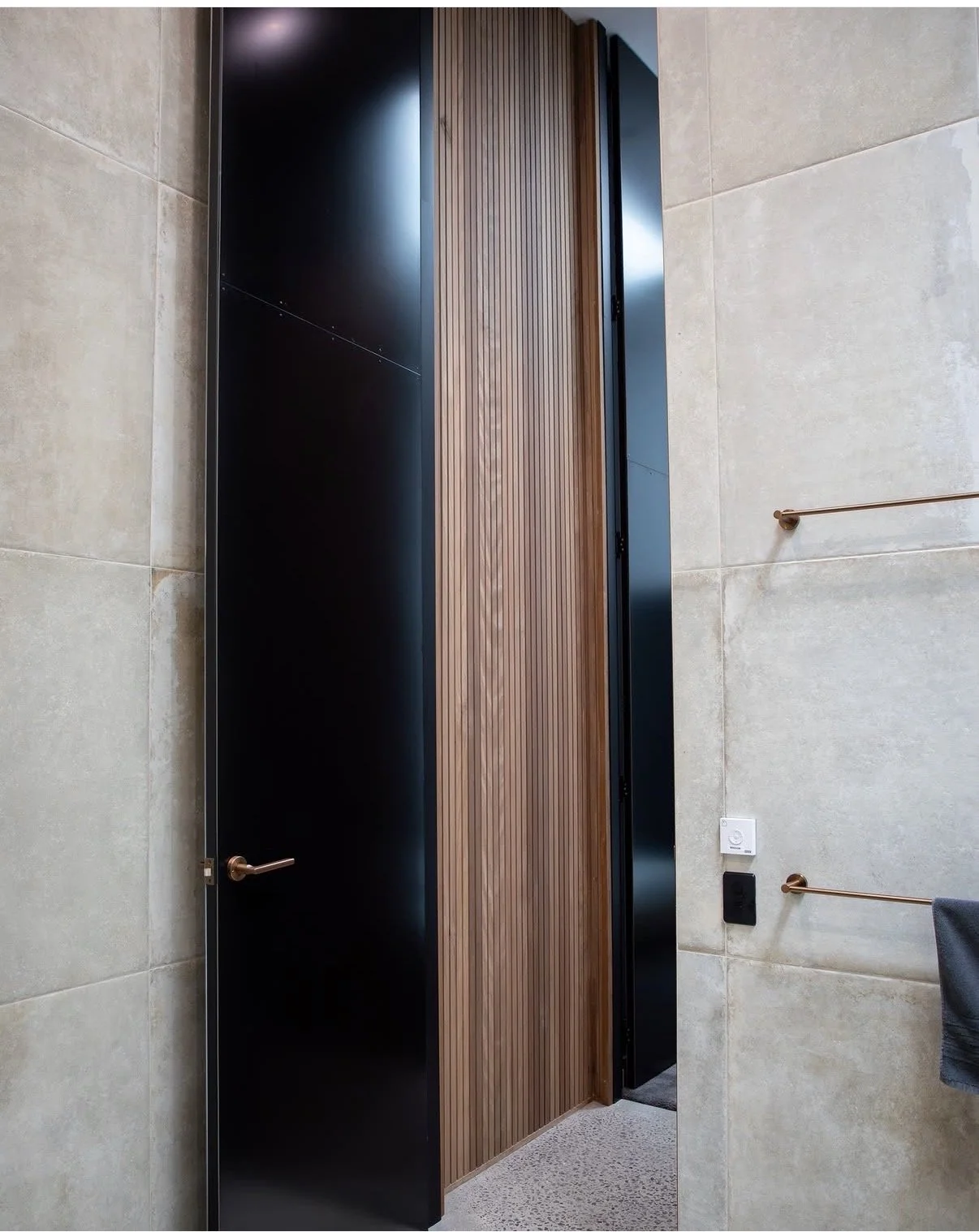 Modern bathroom stall with black door, wooden slats in the middle, beige tiled walls, a towel hanging on a copper towel bar, a switch, and an electrical outlet.