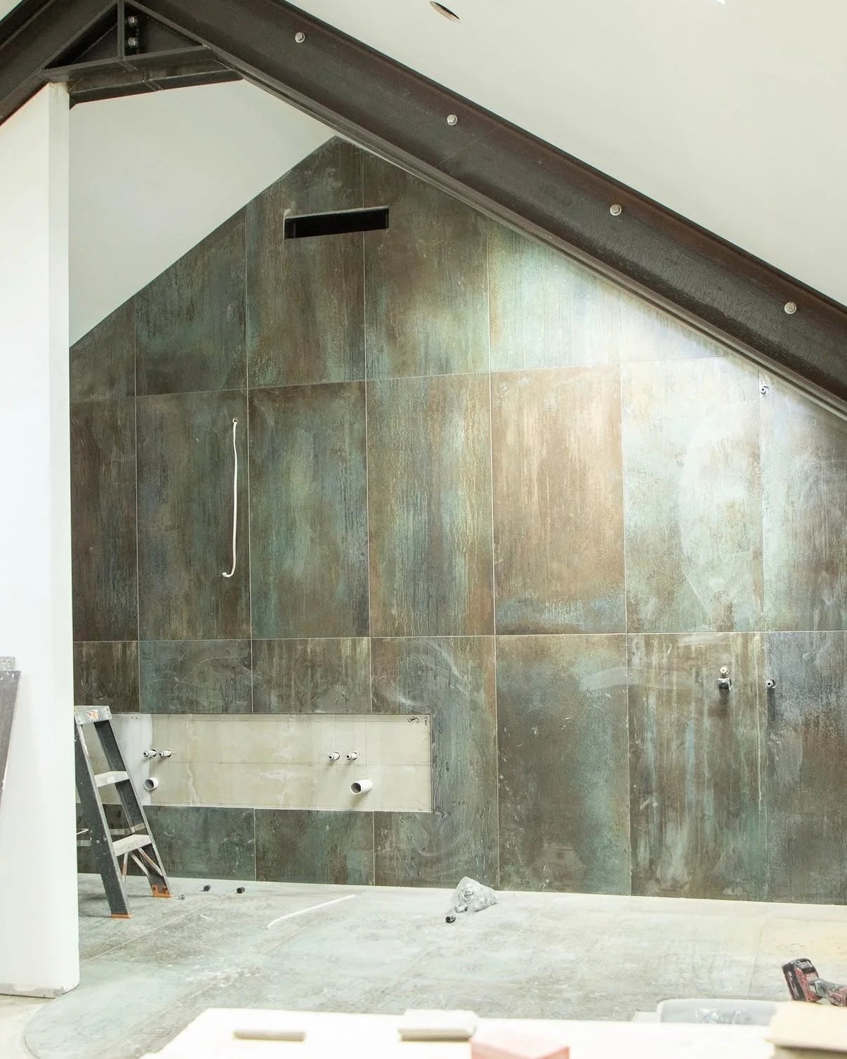 Interior of a room under construction with a sloped ceiling, tiled wall, a ladder, and plumbing fixtures.