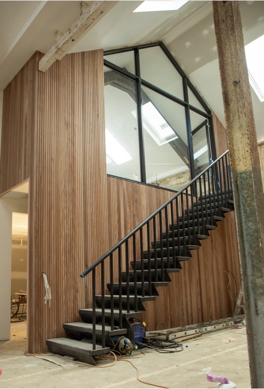 Interior view of a modern staircase with black steps and black railing next to a wooden wall with vertical panels. The area appears to be under construction or renovation, with construction tools and wires on the floor.