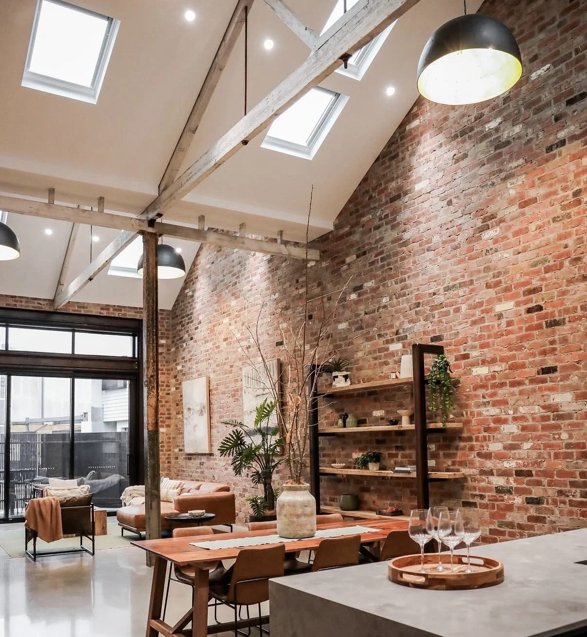Modern living space with exposed brick walls, large windows, and skylights, featuring a dining area with a large vase, a wooden table, and wine glasses, along with a cozy seating area with armchairs and a sofa.