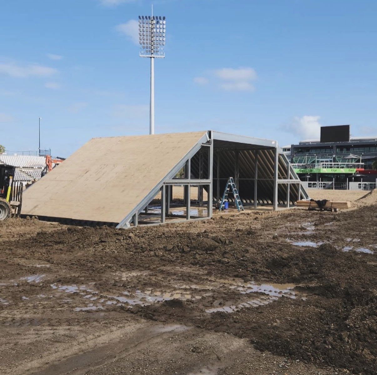 Construction site with a partially assembled building frame and a large sloped wooden roof. A ladder and construction tools are visible on site, and there is a stadium with bright lights and seating in the background under a blue sky.