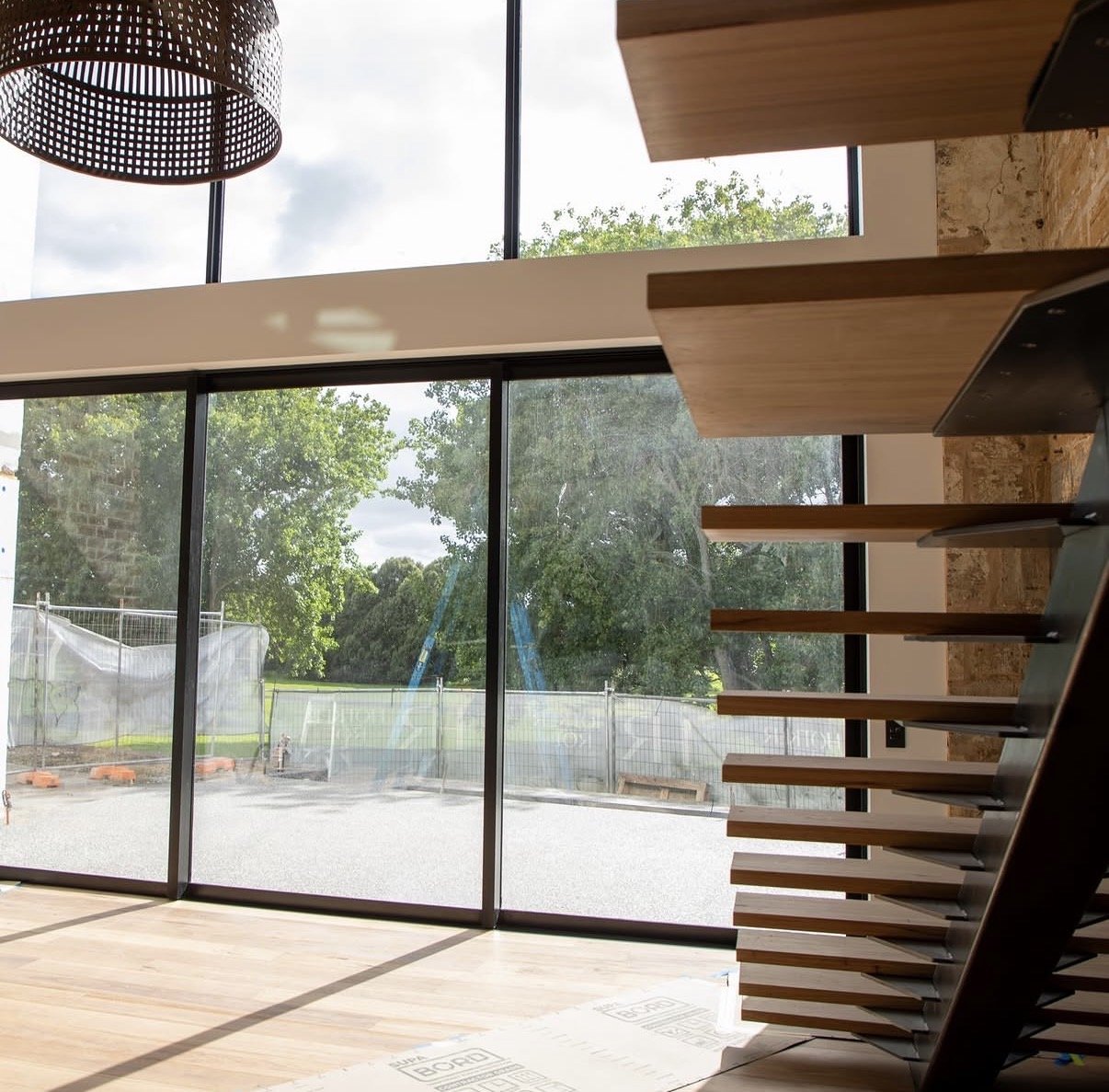 Interior view of a room with large floor-to-ceiling glass windows showing an outdoor construction site and green trees. The ceiling has a round, brown basket-style light fixture. There are wooden stairs with black supports on the right side of the im