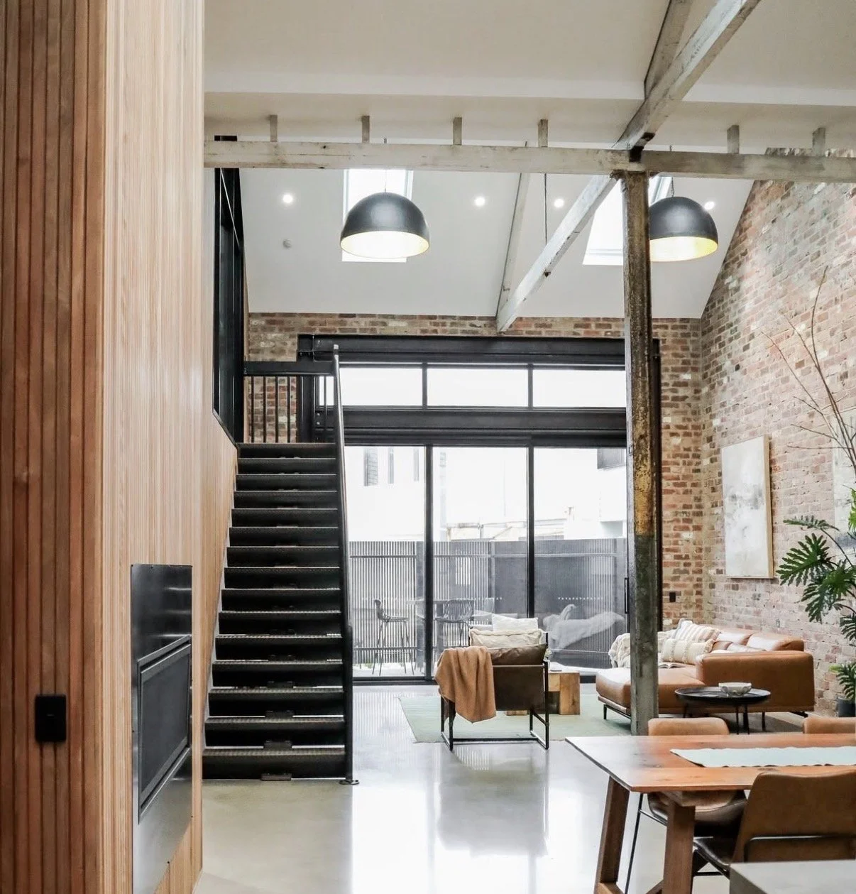 Modern loft apartment with an open living area featuring brick walls, a staircase with black steps, large sliding glass doors to an outdoor patio, contemporary furniture, and ceiling skylights.