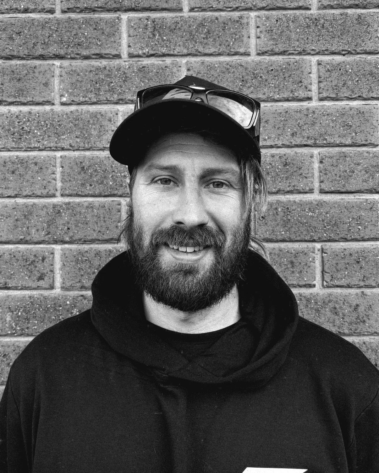 A man with a beard and mustache smiling at the camera, wearing a black hoodie and cap, sunglasses resting on the cap, standing against a brick wall.