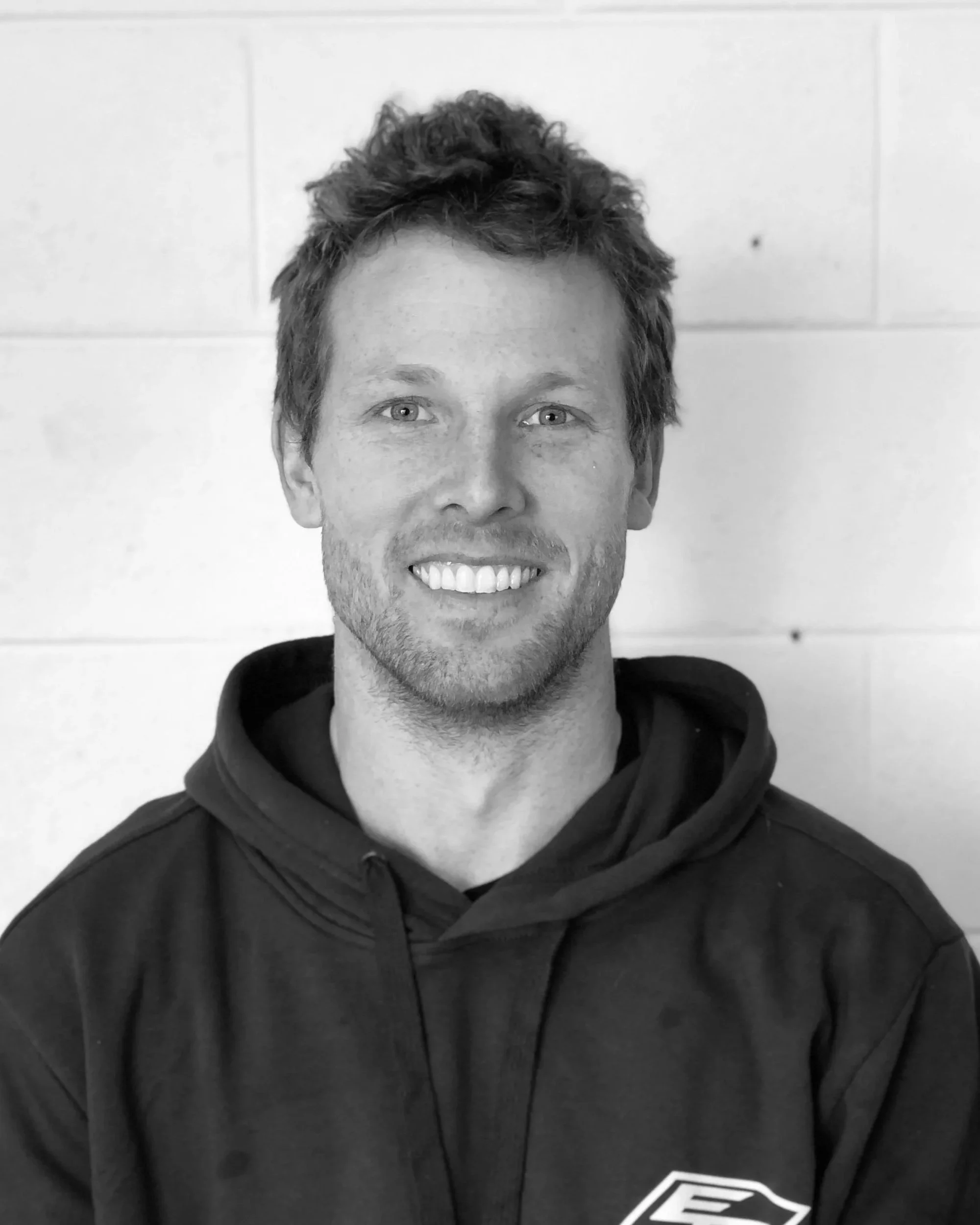 Black and white photo of a smiling man with short, slightly tousled hair, wearing a black hoodie with a logo on the chest, standing against a plain wall.
