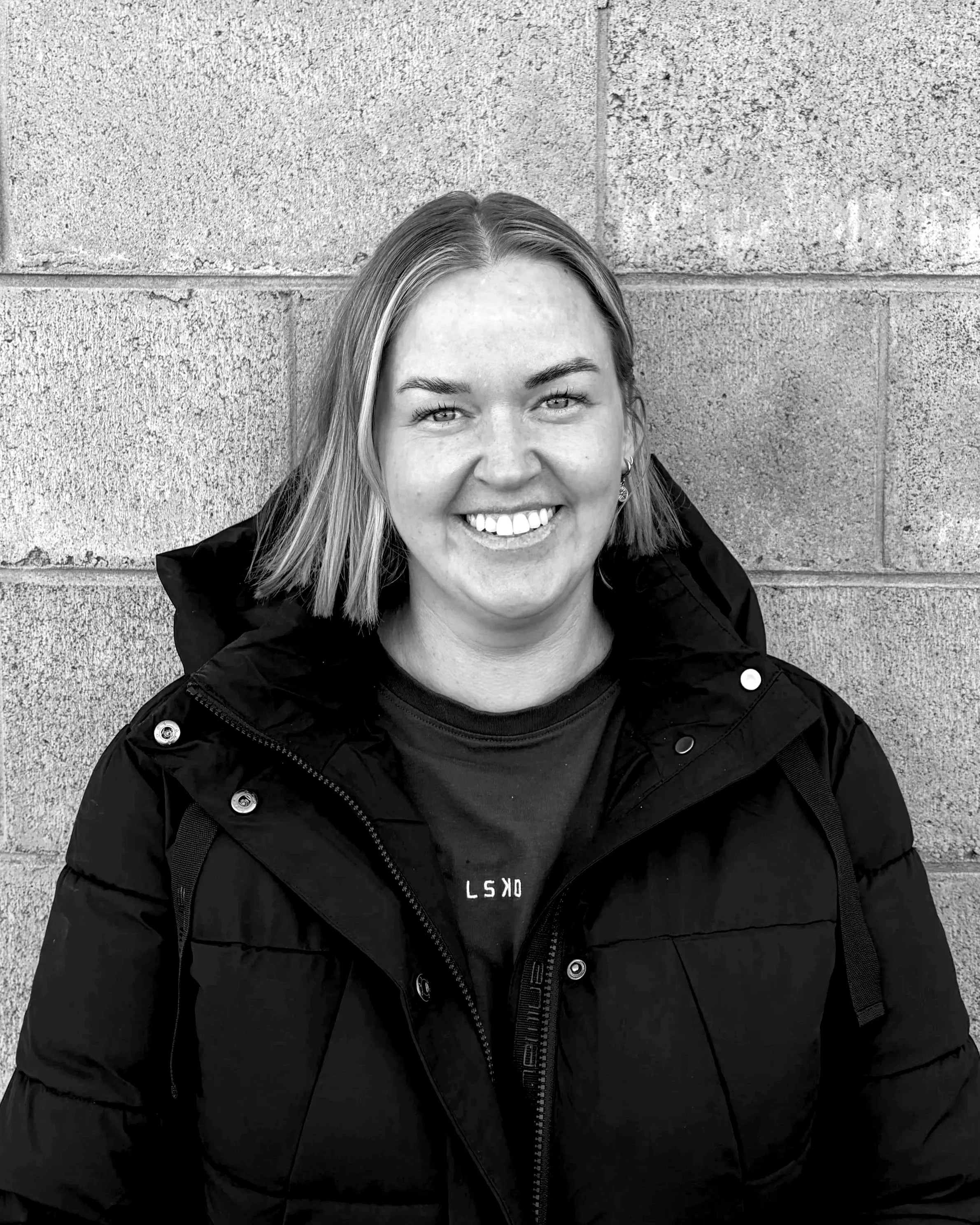 Black and white photo of a woman with light hair, smiling, standing against a brick wall, wearing a dark jacket with zippers and buttons.