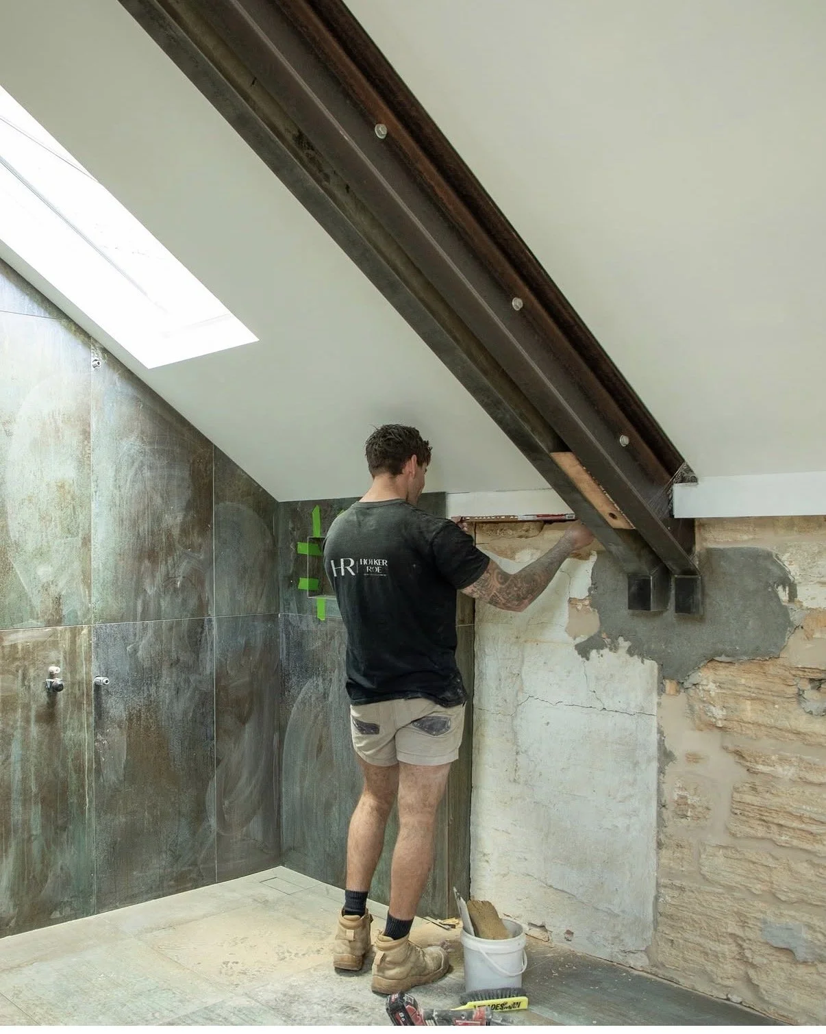 A worker in shorts and a black t-shirt is working on a wall in a room that is under renovation. The room has tiled walls and a sloped ceiling with a skylight, and the worker is using tools near exposed brickwork and concrete.