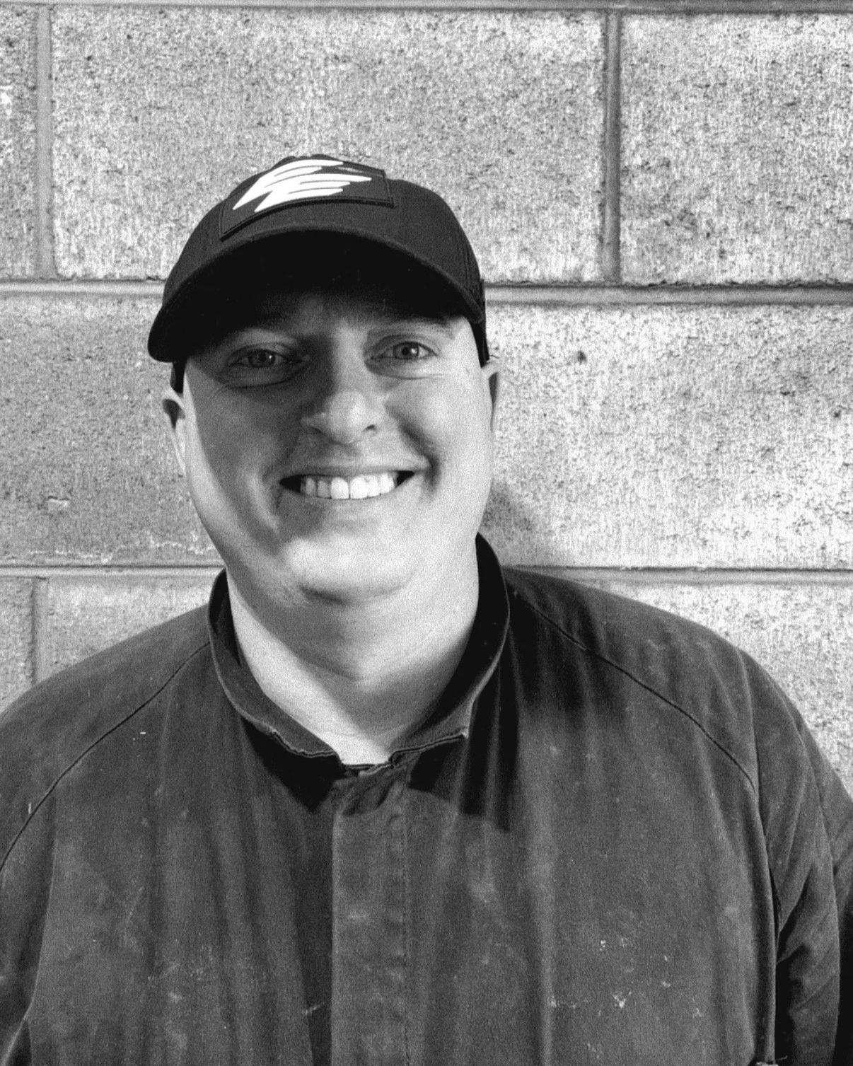 Black and white photo of a smiling man wearing a baseball cap and a jacket, standing against a brick wall.