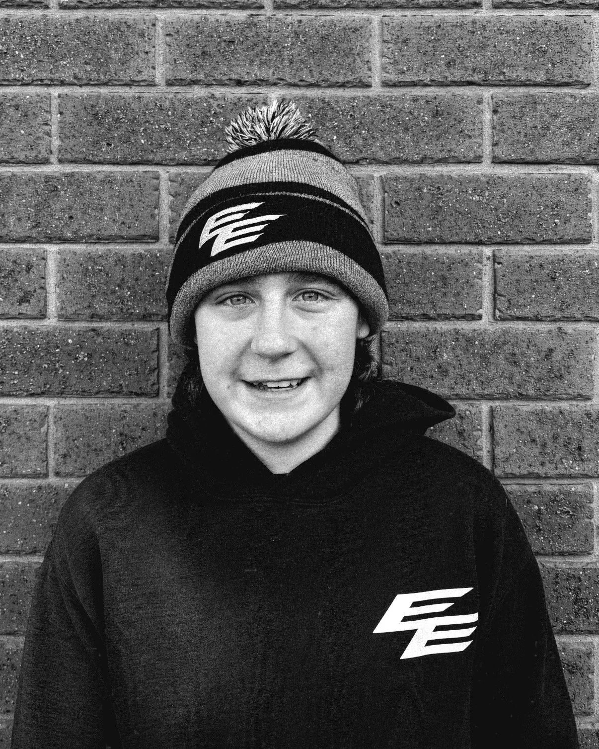 A young person smiling, wearing a knit cap with a pom-pom and a logo, standing in front of a brick wall.