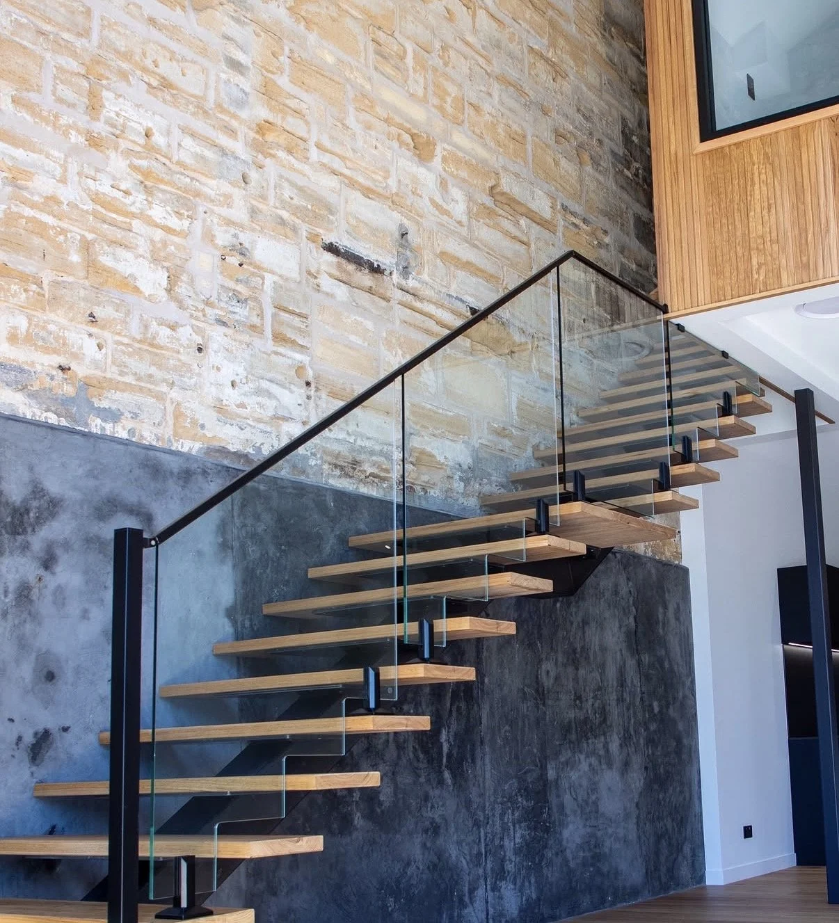 Modern staircase with wood treads, glass railing, black metal supports, against a textured brick wall and concrete wall in a contemporary interior.