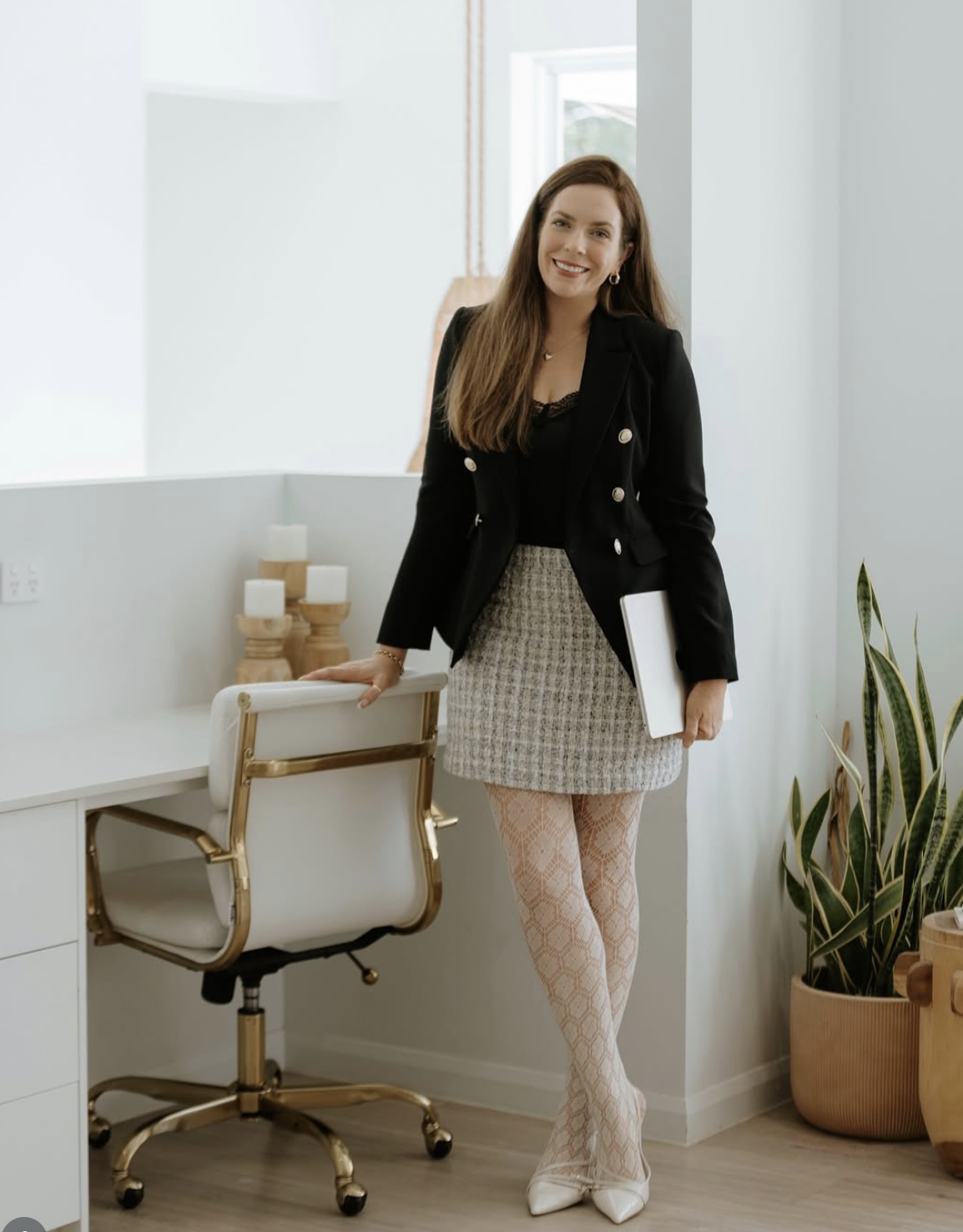 A woman standing in a modern, bright office, smiling, holding a closed laptop, with a stylish white and gold office chair, a desk, candles, and potted plants around her.
