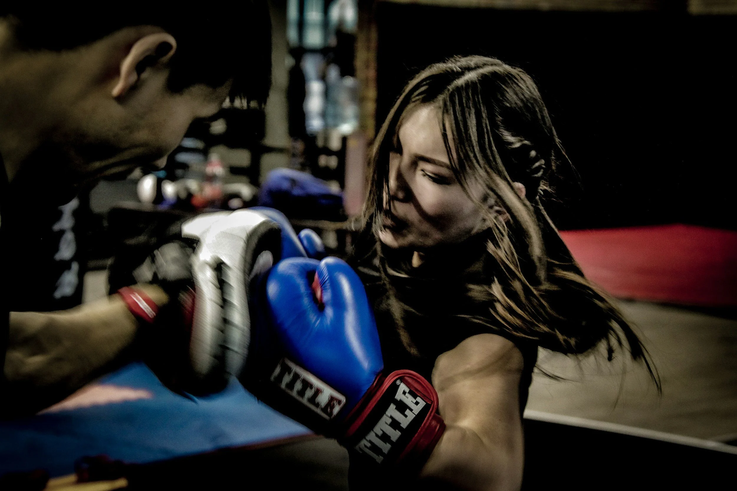 A young female boxer in a gym is practicing boxing with a trainer. She is wearing blue boxing gloves and focusing, with her hair wet and falling over her face. The trainer is holding focus mitts, guiding her punch.
