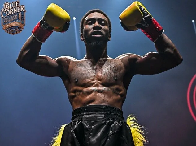 A muscular male boxer with yellow boxing gloves and black shorts stands in a boxing ring, raising his arms in victory.