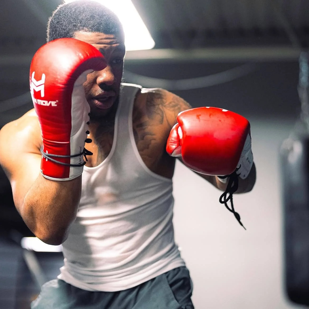 A boxer in a white tank top and grey shorts practicing punches in a gym, wearing red boxing gloves.