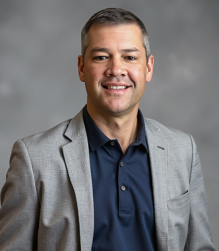 Professional headshot of a middle-aged man with short gray hair, wearing a dark blue collared shirt and a light gray blazer, smiling against a neutral background.