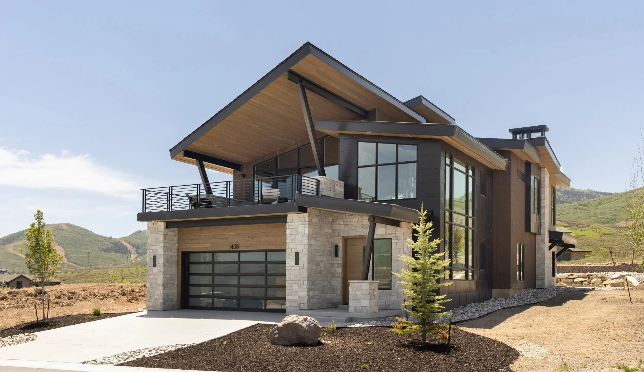 Modern house with large glass windows and a balcony, set against a landscape of rolling hills under a clear sky.