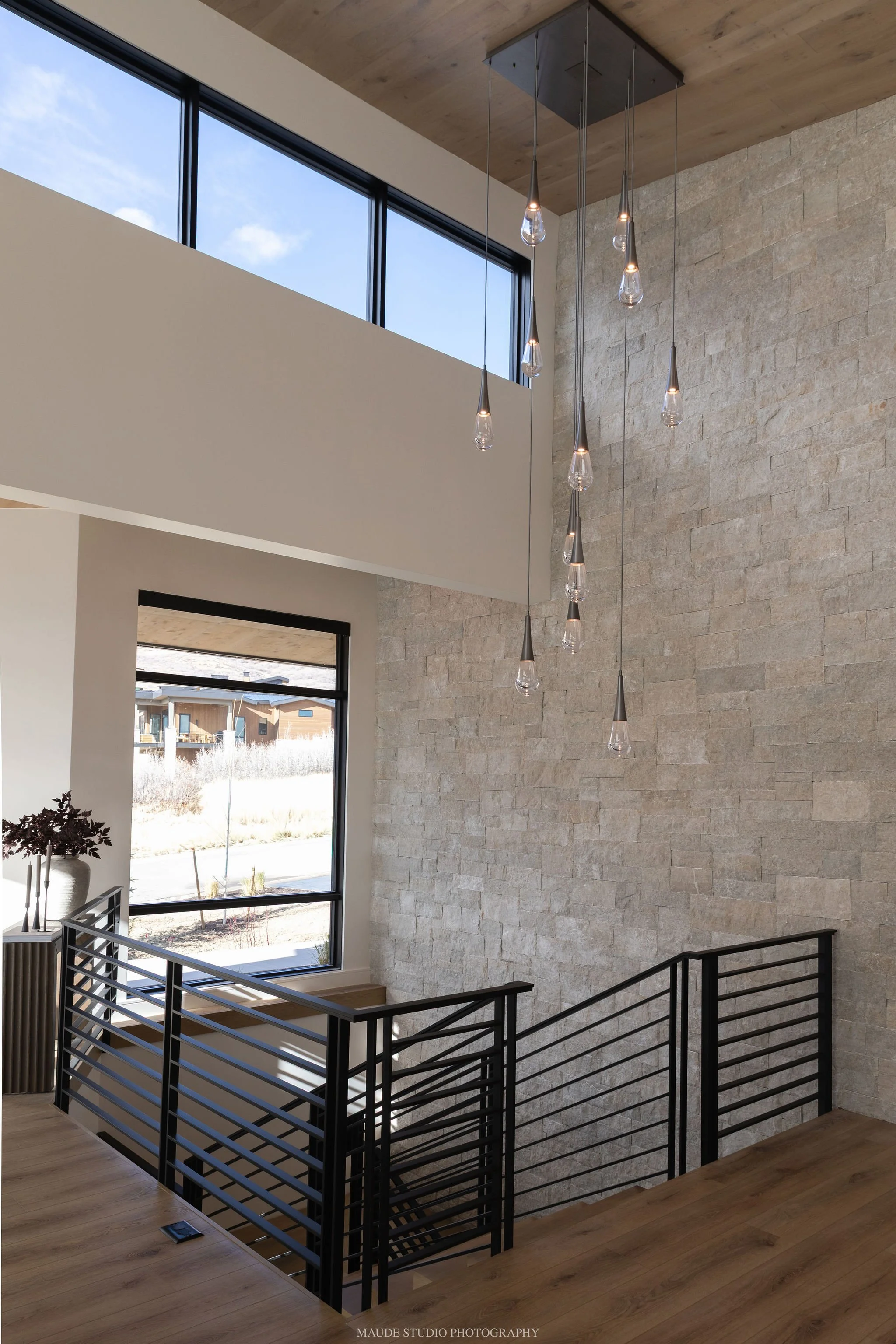 Inside view of a modern house staircase with black metal railings and light wood flooring. Large windows letting in natural light, high ceiling with hanging light fixtures, and a textured stone wall.