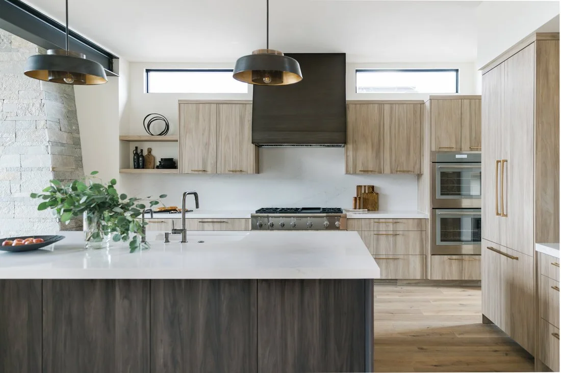 Modern kitchen with light wood cabinets, white marble countertop, black range hood, stainless steel oven, and large island with a plant and a bowl of tomatoes.
