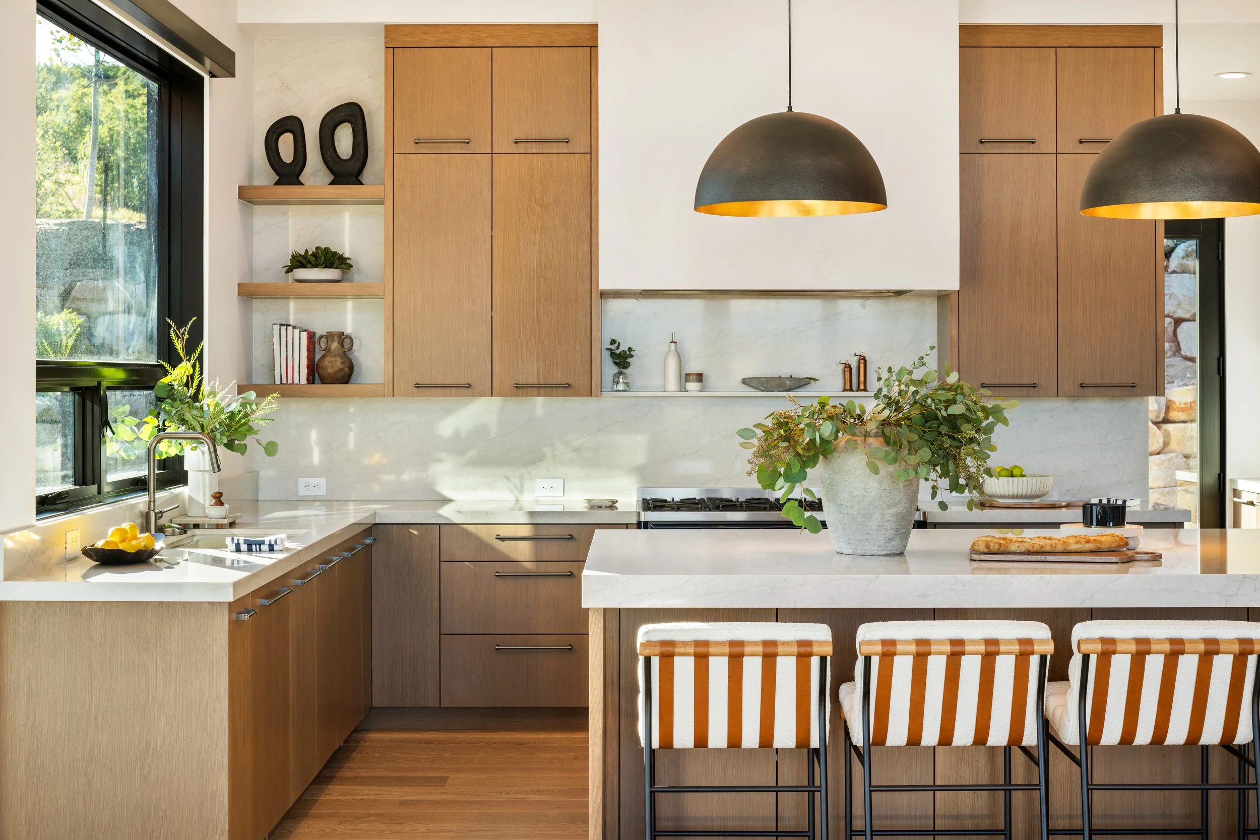 Modern kitchen with wooden cabinets, a large window, pendant lights, and a kitchen island with a plant and bar stools.