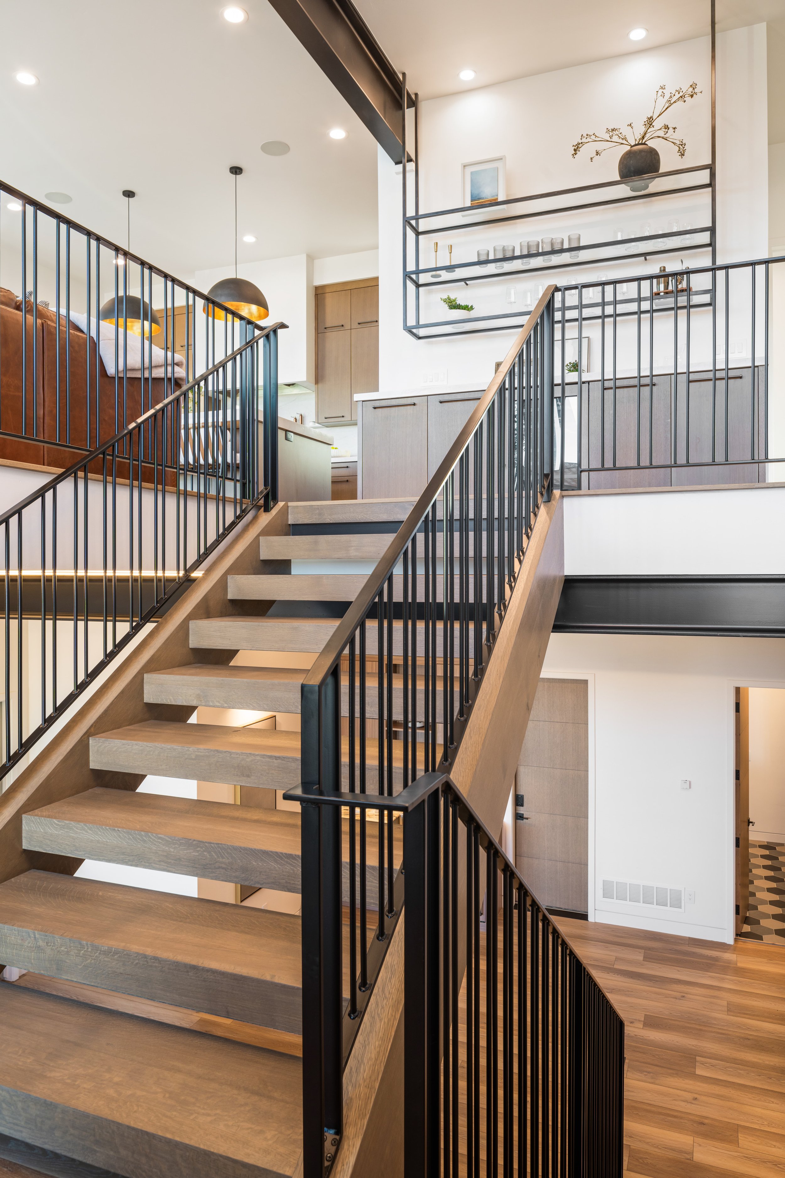 Modern staircase with wooden steps, metal railing, and open risers in a contemporary home with a second-floor balcony, kitchen in background, and ceiling lights.