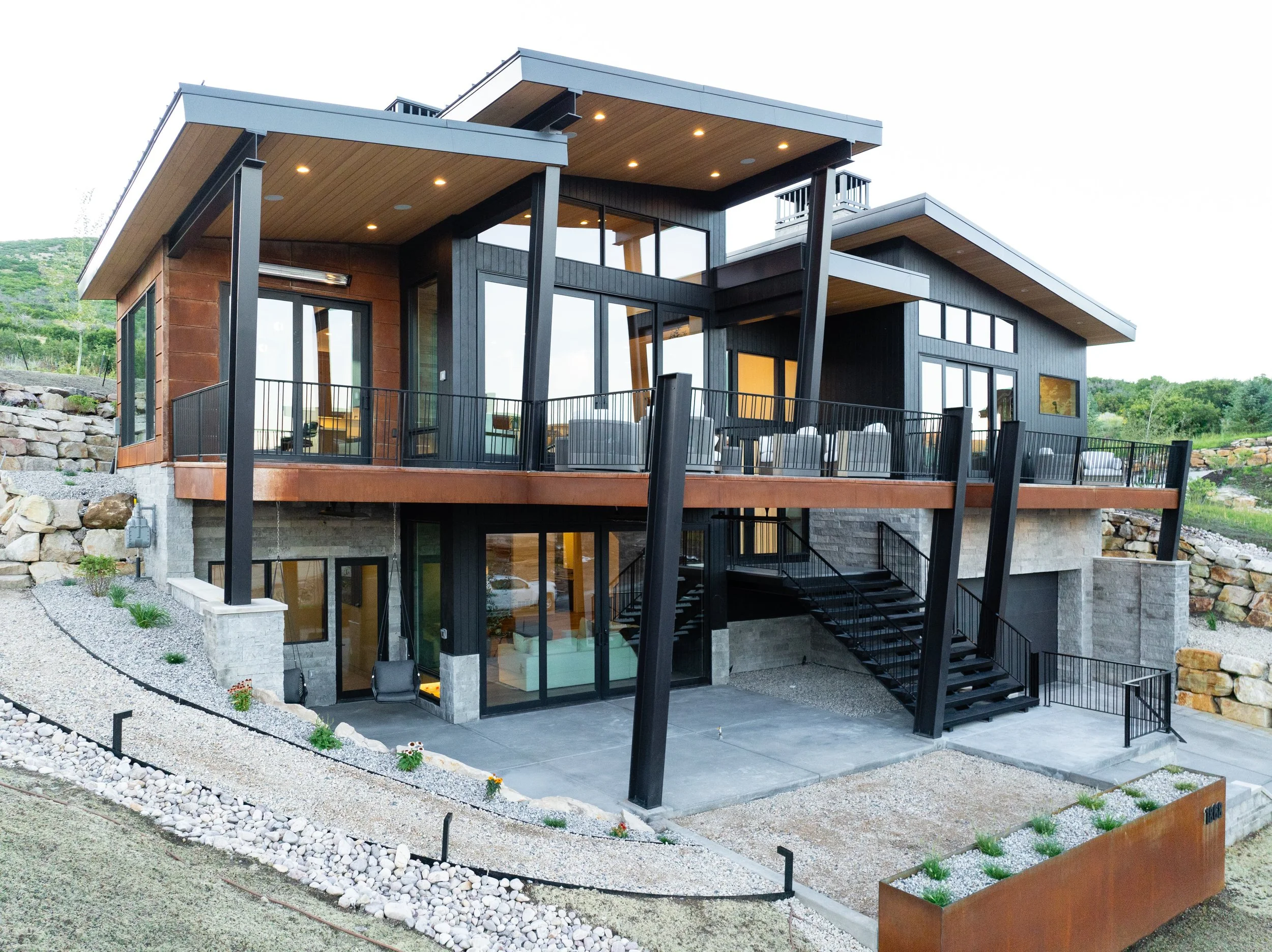Modern multi-level house with large glass windows, black and wooden exterior, outdoor patio, metal staircase, and landscaped yard with rocks and plants.