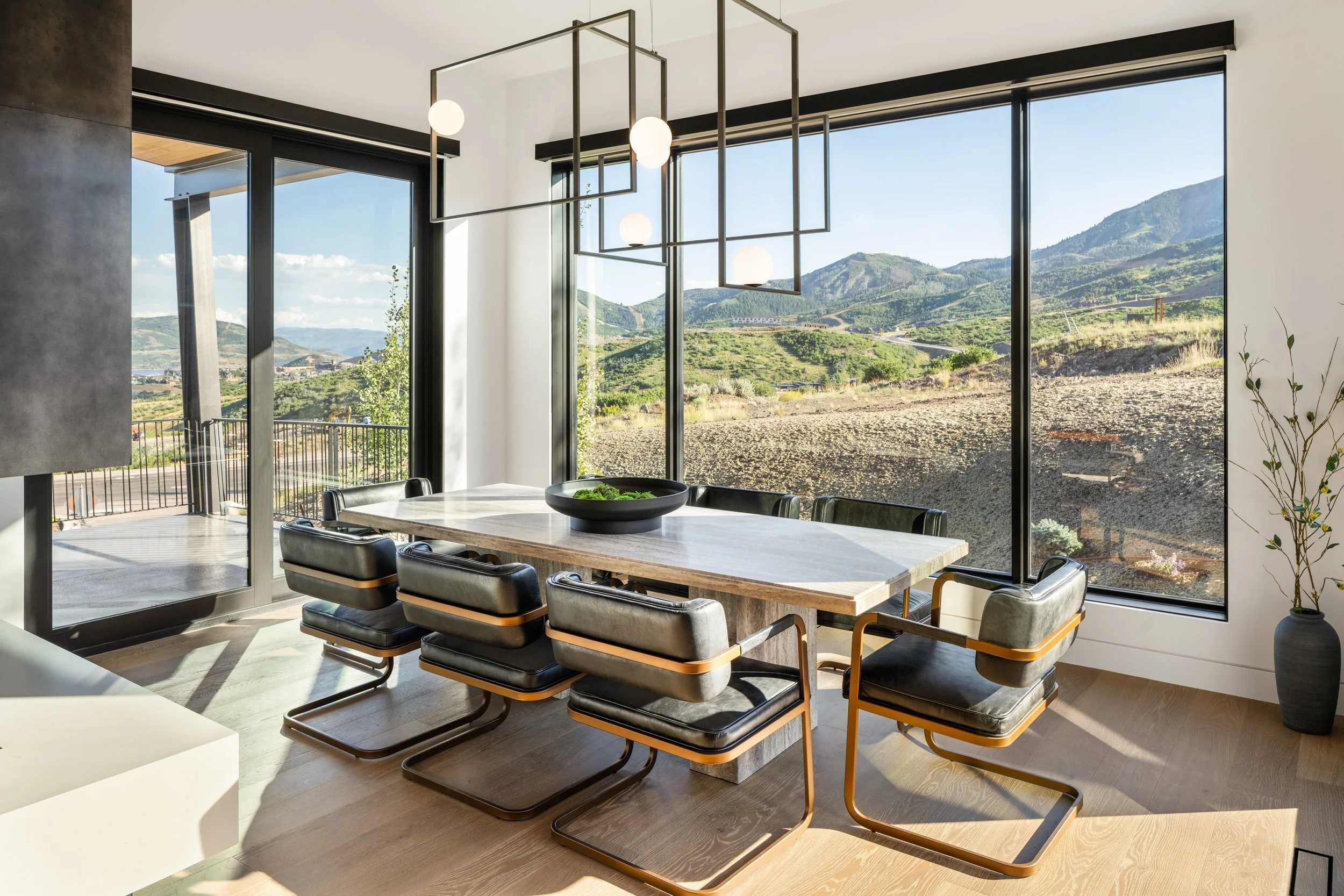 Modern dining room with large floor-to-ceiling windows showing mountainous landscape, minimalist chandelier above marble dining table, black leather chairs, and a small potted plant in the corner.