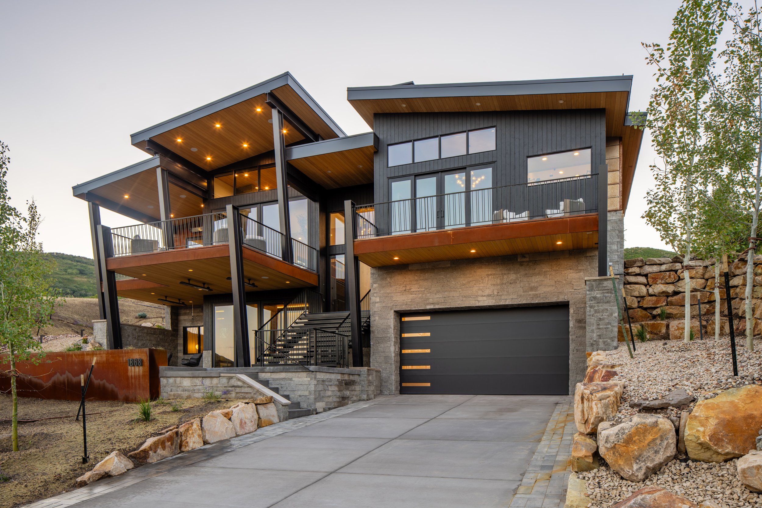 Modern multi-story house with large windows, balconies, and an attached garage, situated on a sloped landscape with rocks and a concrete driveway, during dusk with exterior lights on.