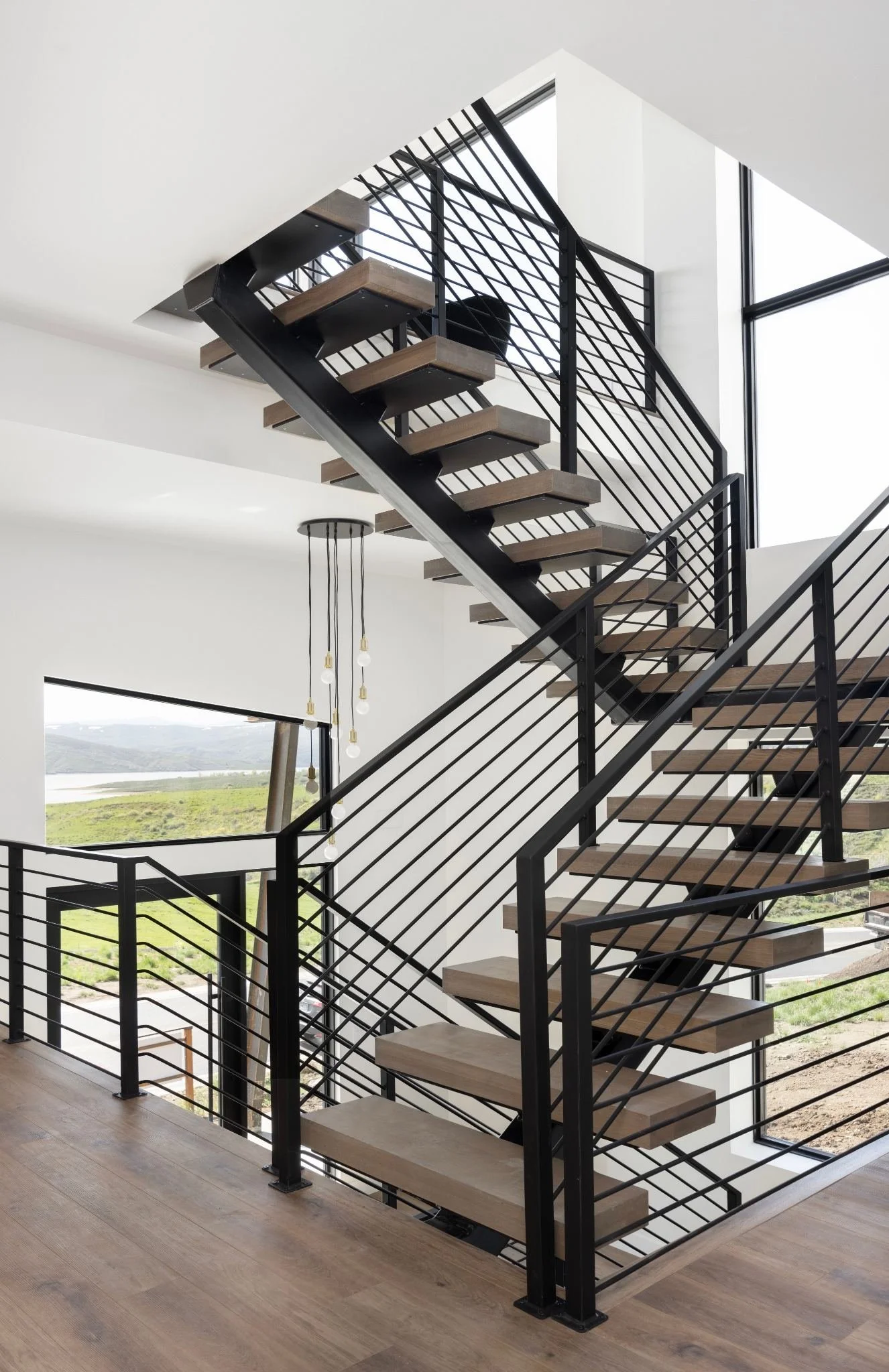 Modern interior staircase with black metal frame, wooden steps, and black metal railing, overlooking a landscape with green fields and a body of water outside large windows.