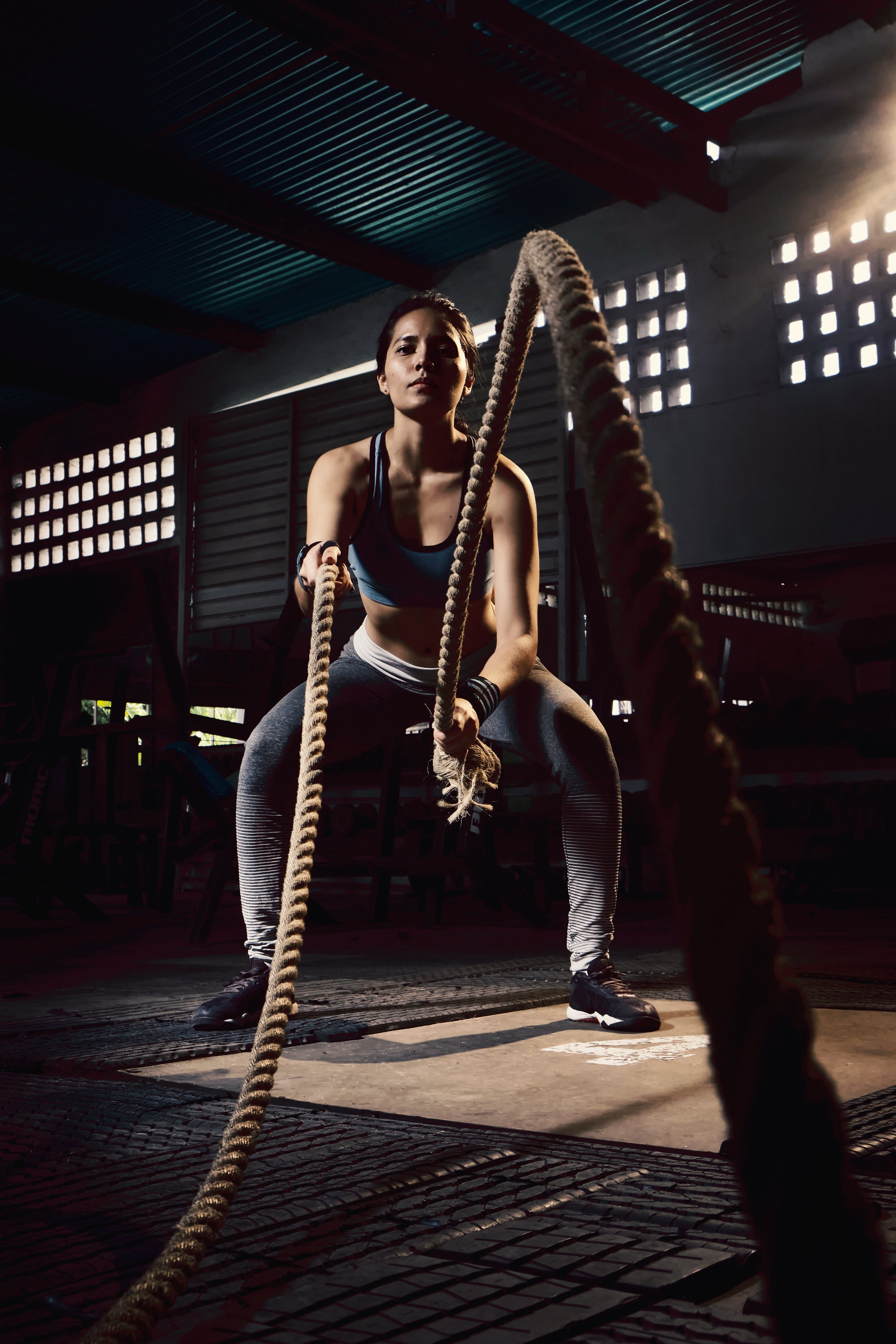 Female athlete in workout attire gripping battle ropes inside a gym with dim lighting and industrial-style windows.