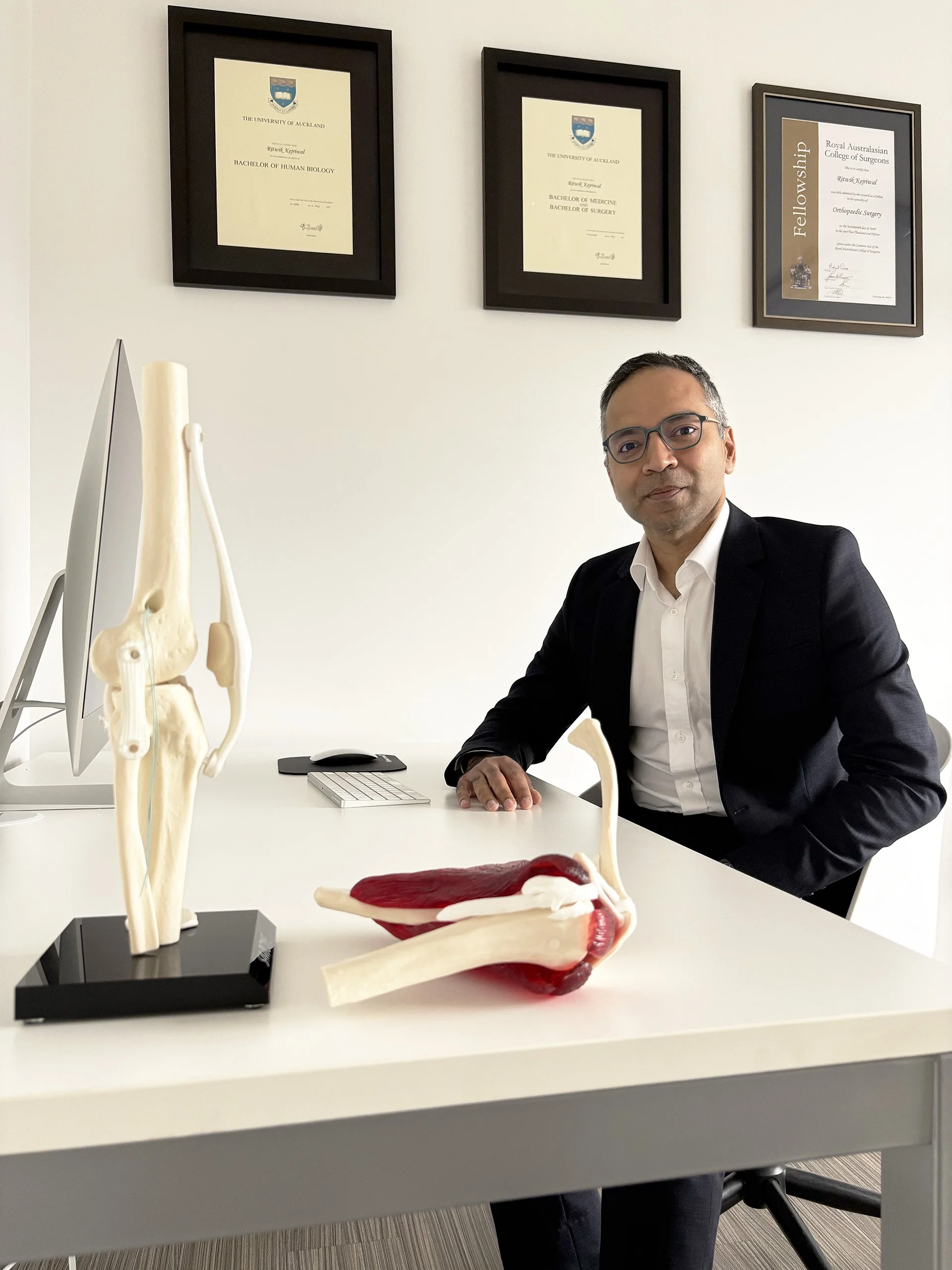 A man in a suit sitting at a desk with anatomical models of bones and ligaments, and framed diplomas on the wall behind him.