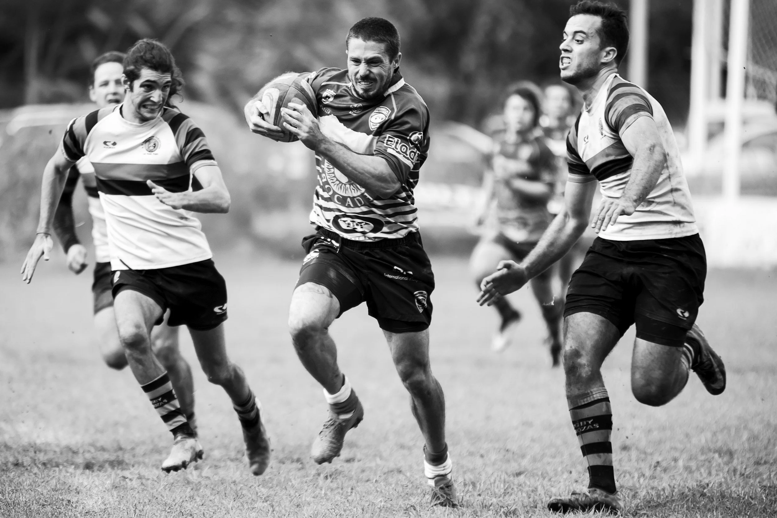 Black and white photo of a rugby game with several players chasing the ball, some with muddy uniforms, on a grass field.