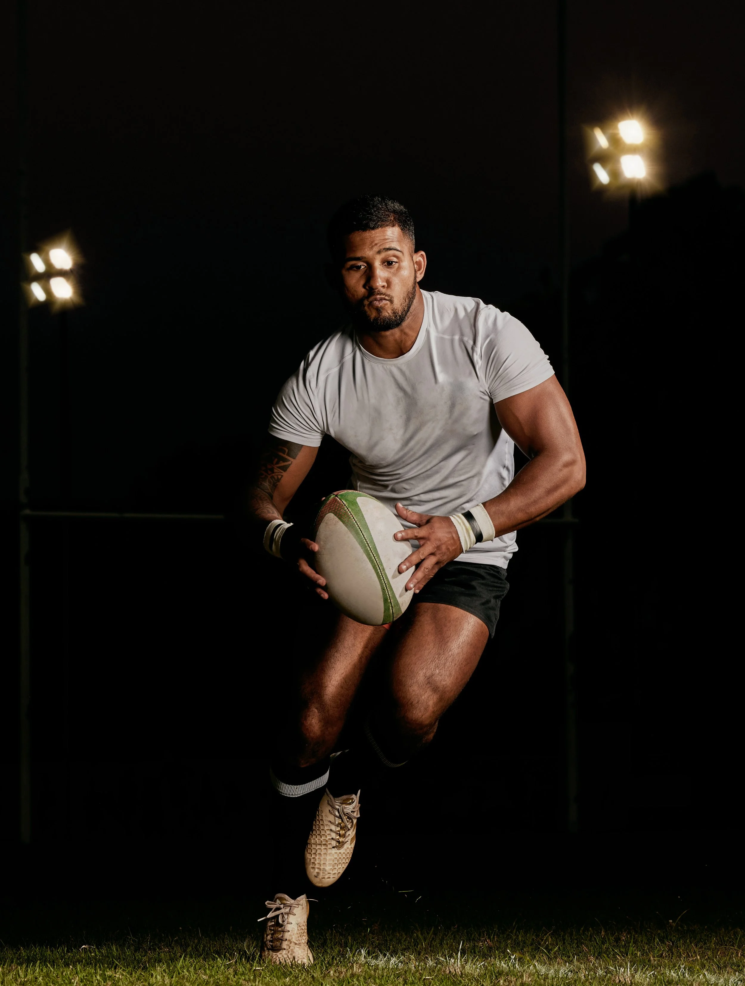 A male rugby player holding a rugby ball, dressed in a white shirt, black shorts, and football shoes, on a grassy field at night with stadium lights.