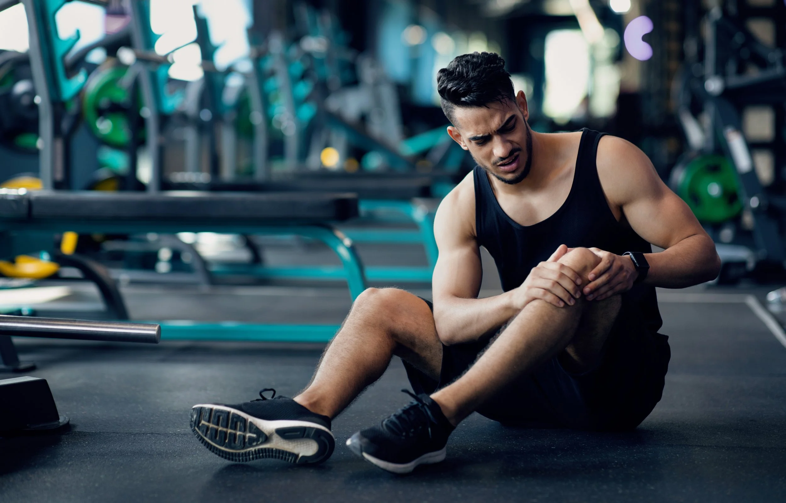 Man in black athletic tank top and shorts sitting on gym floor, holding his ankle, with a pained expression, in a gym with workout equipment in the background.