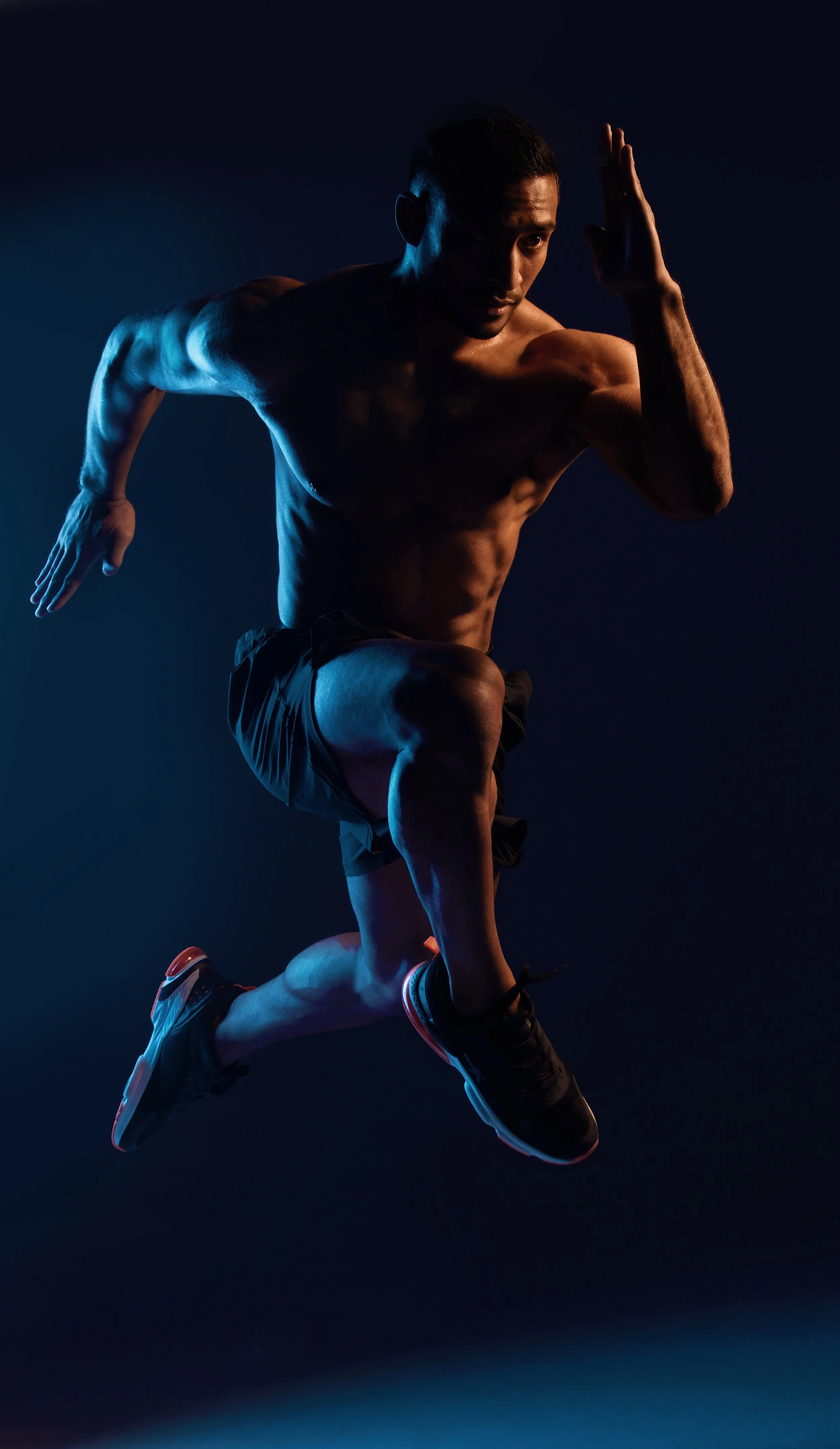 A shirtless man in black shorts and running shoes is posing mid-motion against a dark background, with dramatic lighting highlighting his muscular physique.