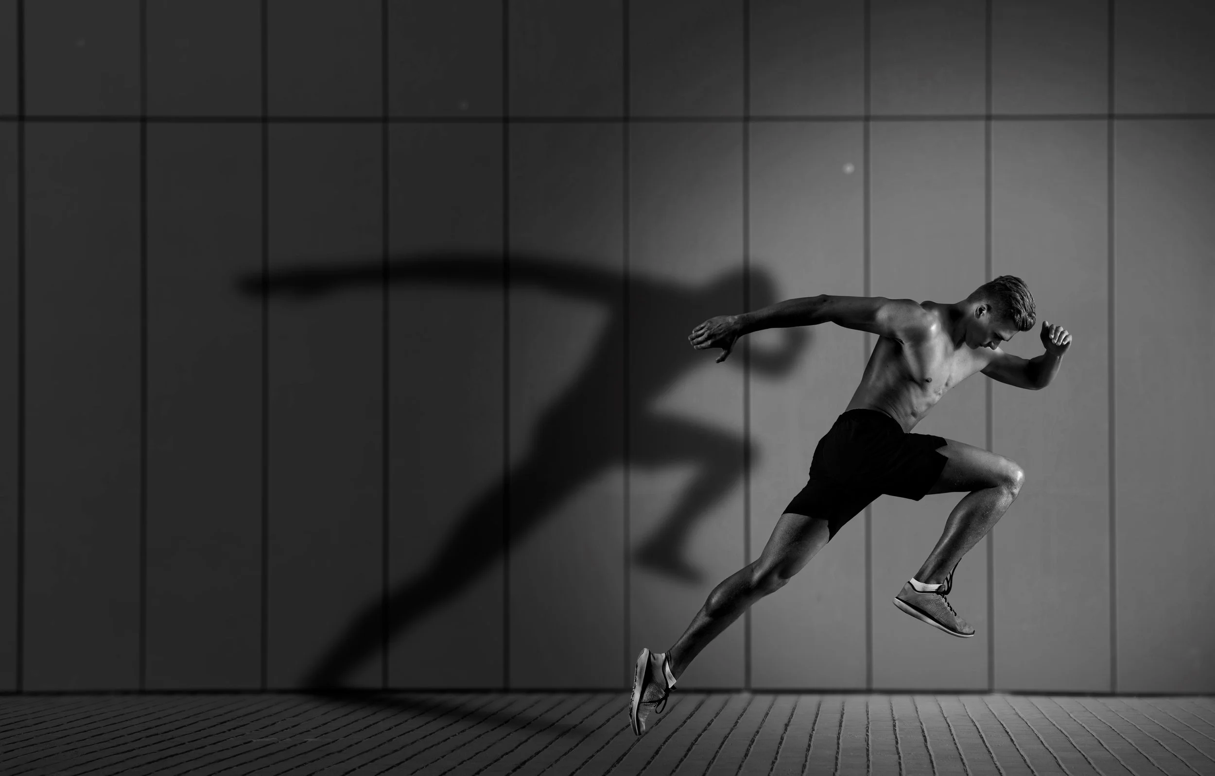 A shirtless male athlete running indoors against a tiled wall, casting a shadow.
