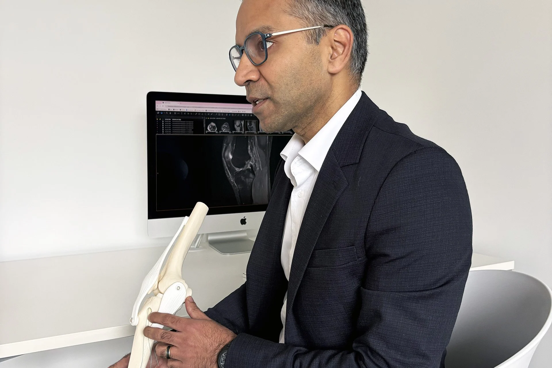 A man in a suit with glasses examining a human knee joint model while sitting at a white desk, with a computer displaying MRI images of a knee in the background.