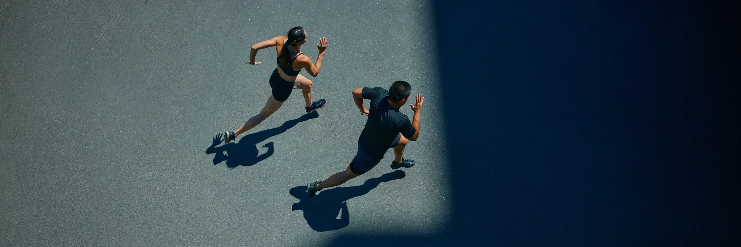 A man and woman running on a paved surface, creating shadows on the ground under a bright light with a dark background.