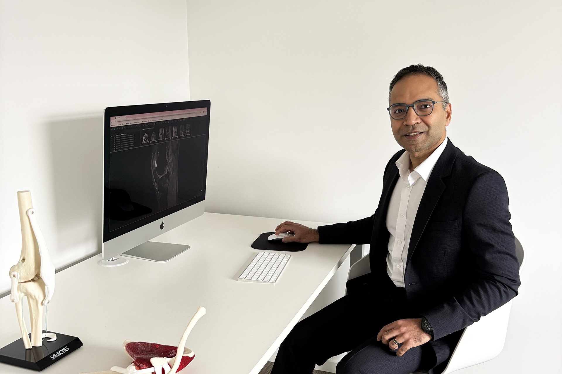 A man in a black suit with glasses sitting at a white desk with a computer, a skeletal spine model, and anatomical pelvis model in a well-lit office with white walls.