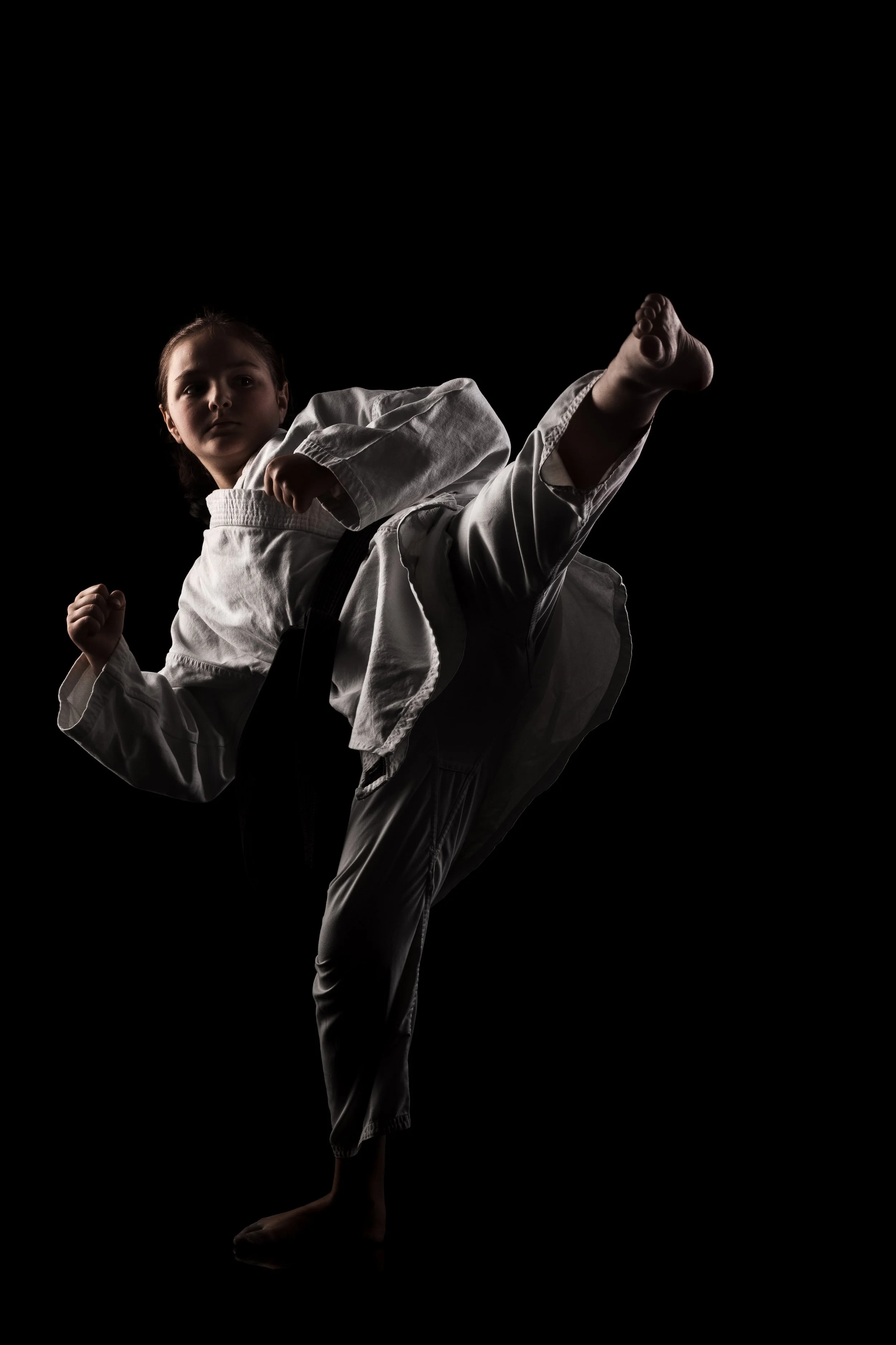 A young girl dressed in a white martial arts uniform performs a high kick against a black background.
