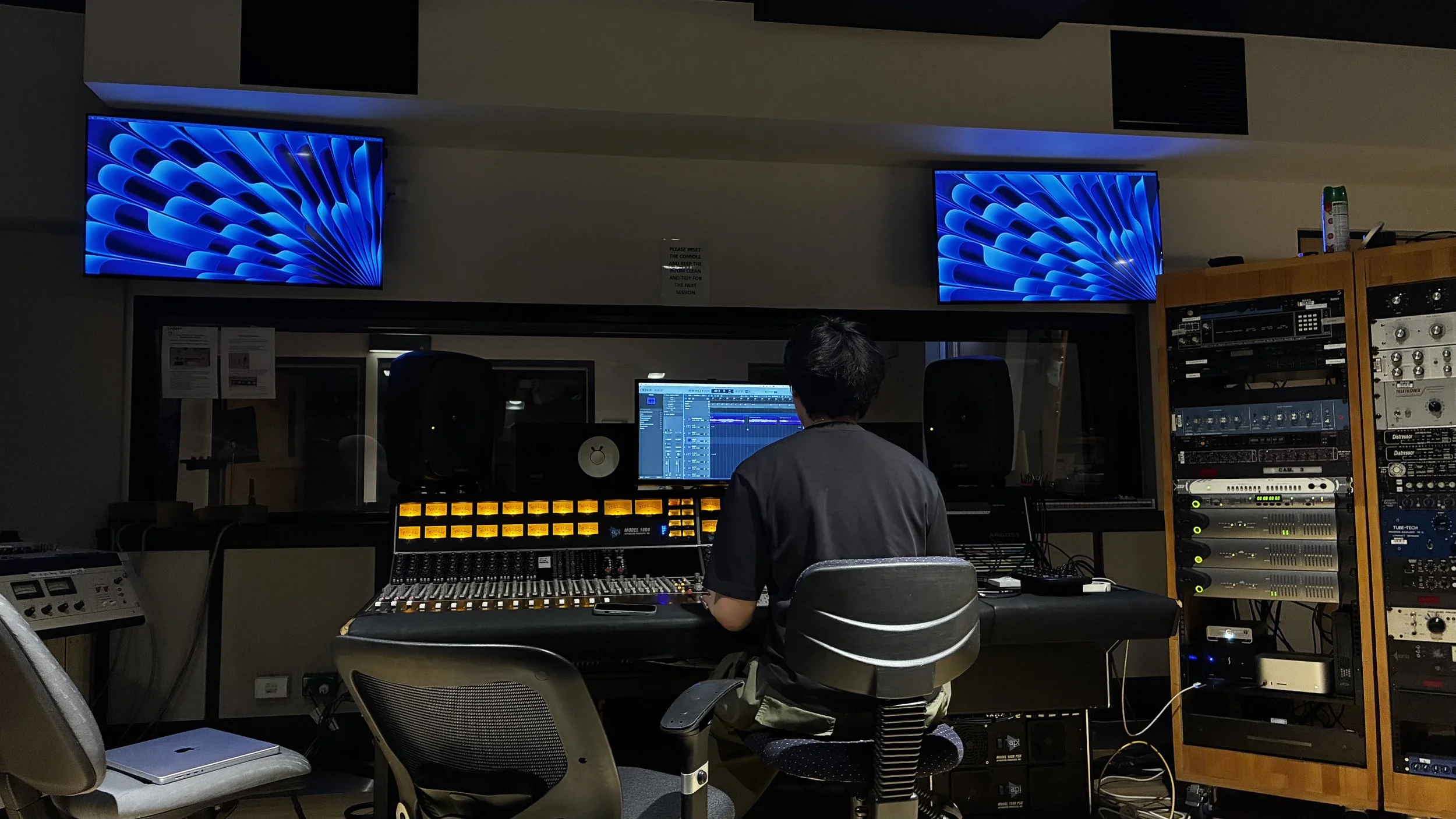 A person working in a recording studio wearing headphones, seated at a mixing console with multiple monitors, surrounded by audio equipment and large screens displaying abstract blue patterns.