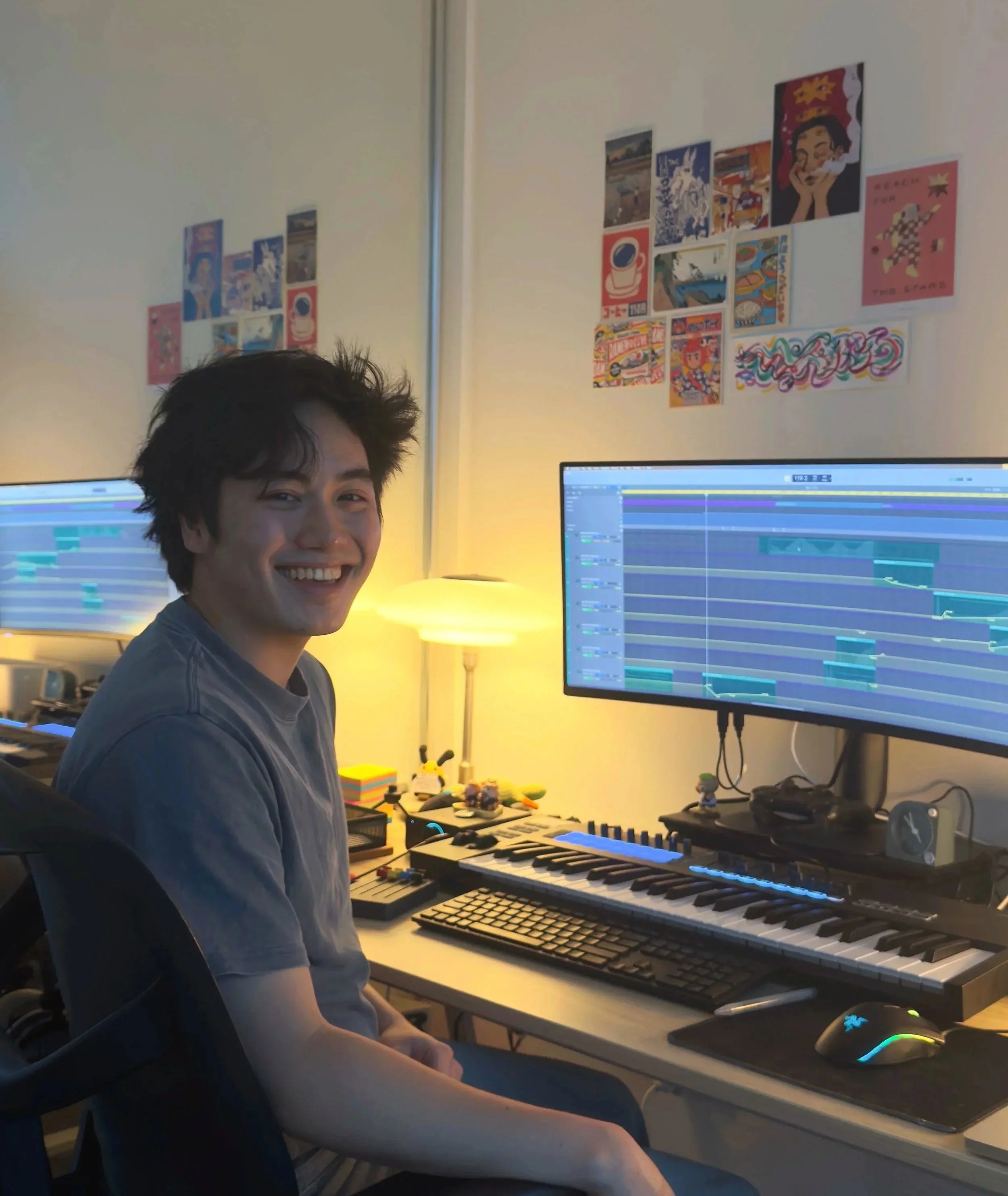 A young man sitting at a home music production setup, smiling at the camera. He is in front of a computer monitor displaying music editing software, with a keyboard, MIDI controller, and mouse on the desk. The background wall has various colorful posters and artwork.