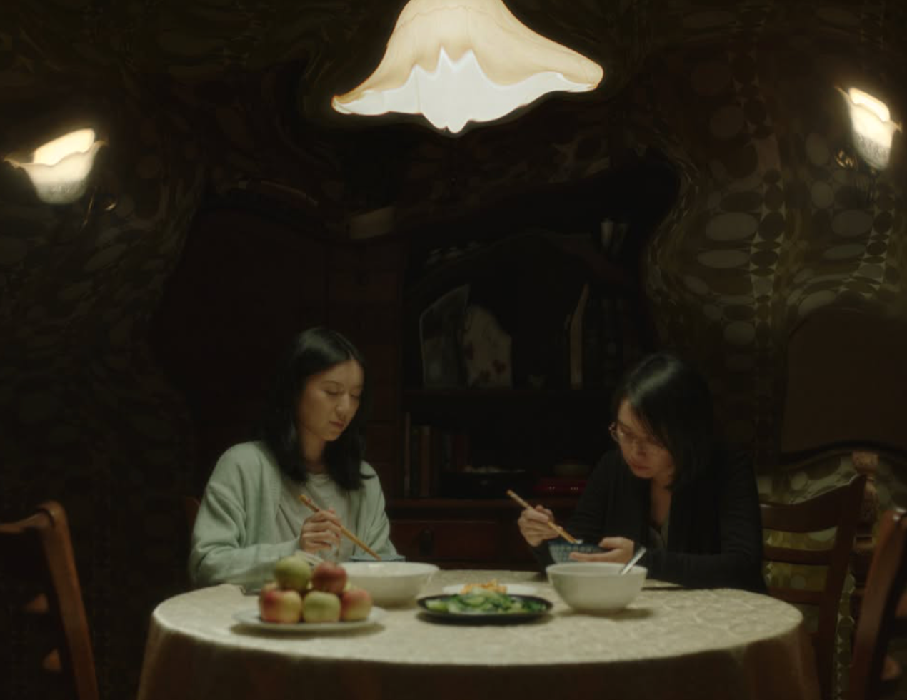 Two women sitting at a round dining table with bowls, a plate of apples, and vegetables in a dimly lit room, eating with chopsticks.