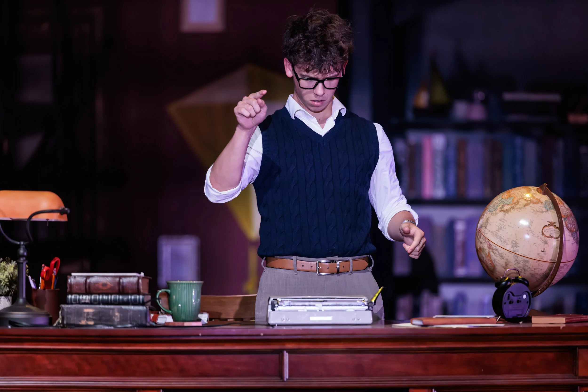 A young man with curly hair and glasses wears a white shirt and a navy sweater vest, standing behind a desk with various office supplies, a globe, and a typewriter in front of him, in a room with bookshelves in the background.