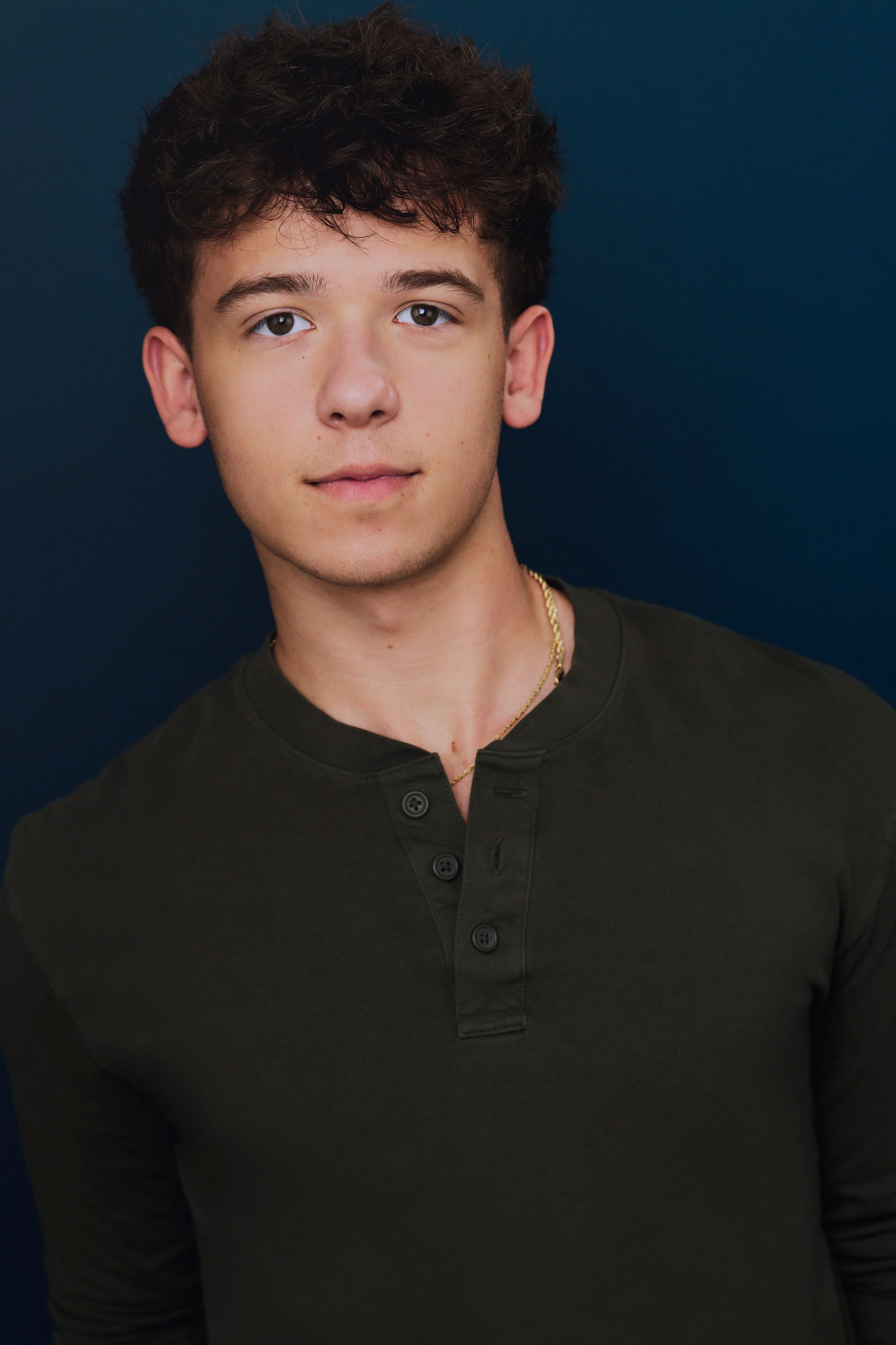 A young man with brown, curly hair and light skin, wearing a dark green buttoned shirt and a gold chain necklace, looking directly at the camera against a dark blue background.