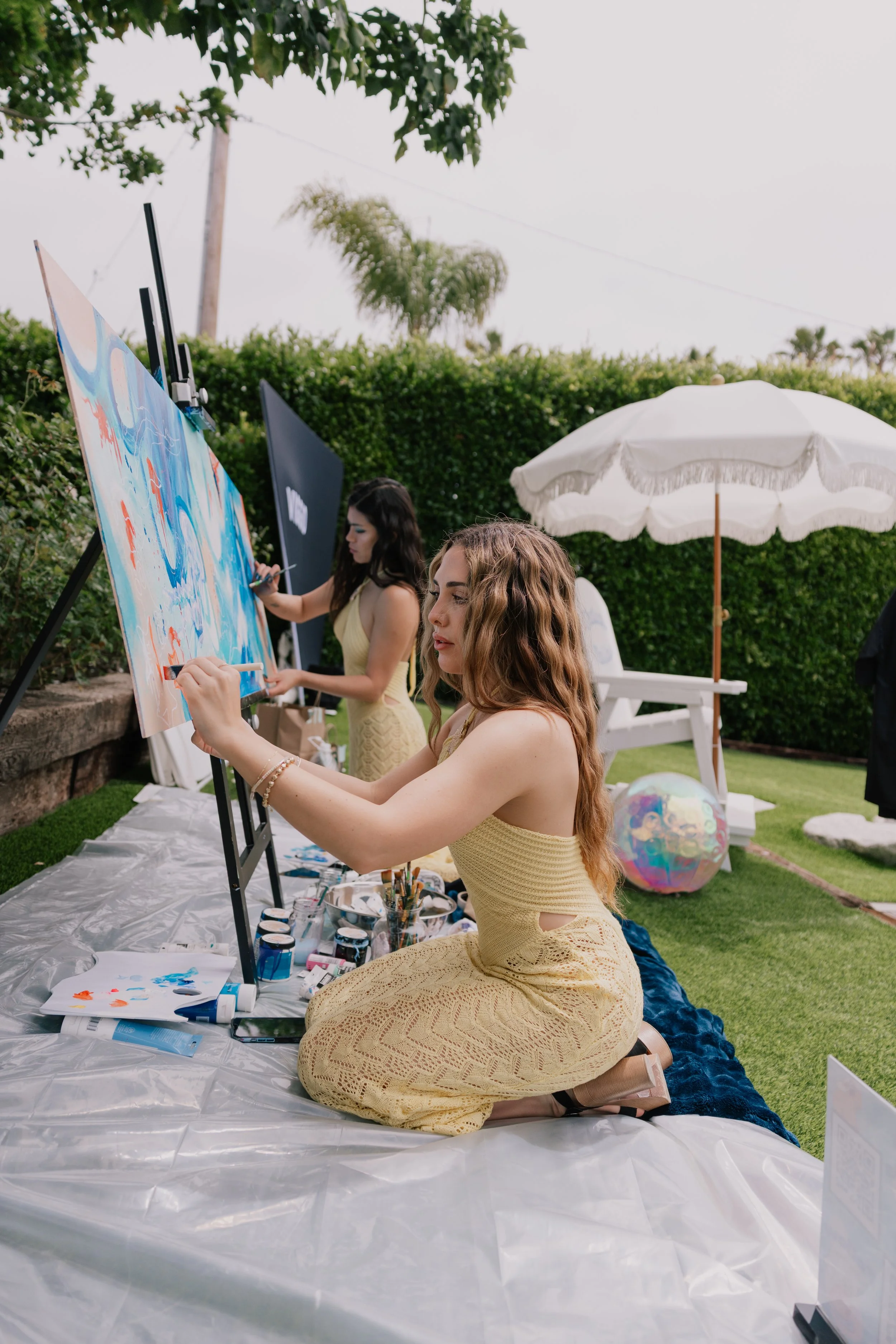 Two women with long, wavy hair in yellow dresses kneeling on a white tarp, painting at an outdoor art event with a canvas, brushes, and paint on the ground. There are umbrellas and a rainbow beach ball in the background.